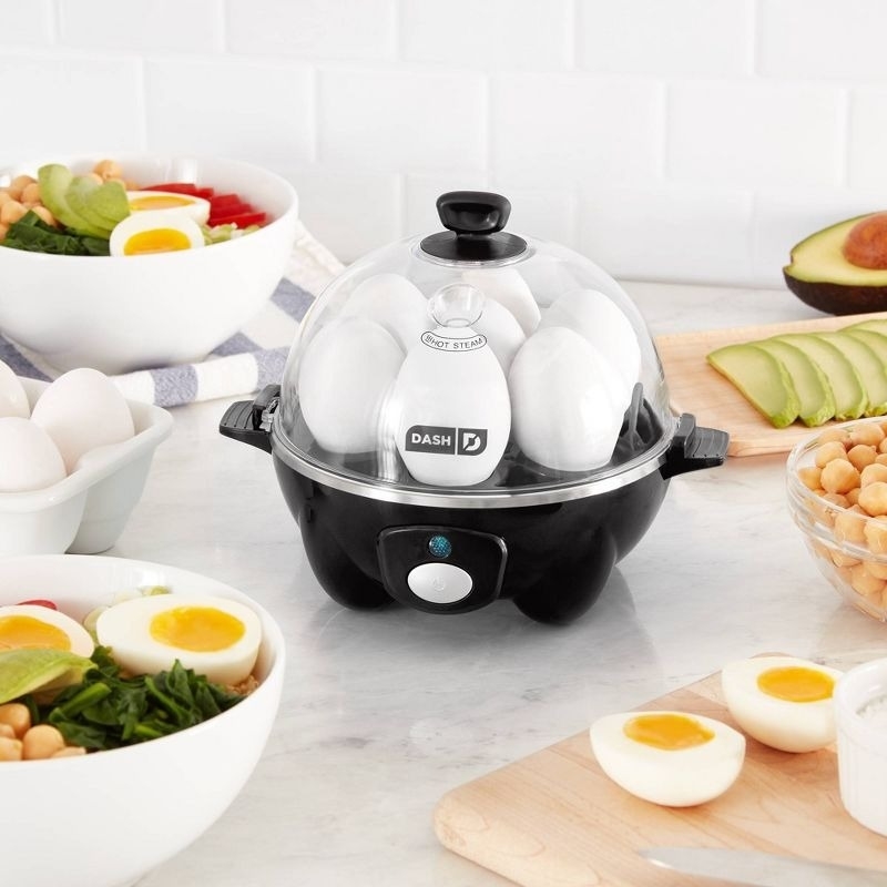 A Dash egg cooker is on a kitchen counter surrounded by bowls of salads and chopped vegetables, with some hard-boiled eggs