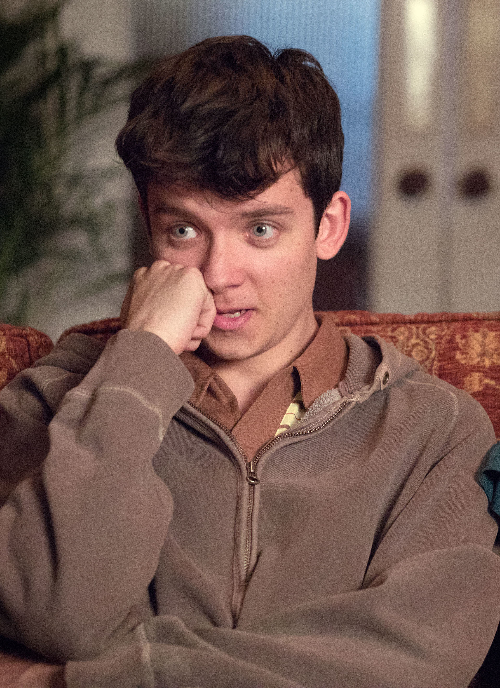 Asa Butterfield sitting on a couch, wearing a zipped-up hoodie, resting his head on his hand, with a pensive expression