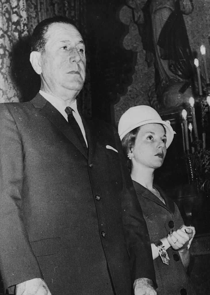 A man in a suit and a woman wearing a hat and a suit stand together in what appears to be a formal setting. Names of the individuals are not known