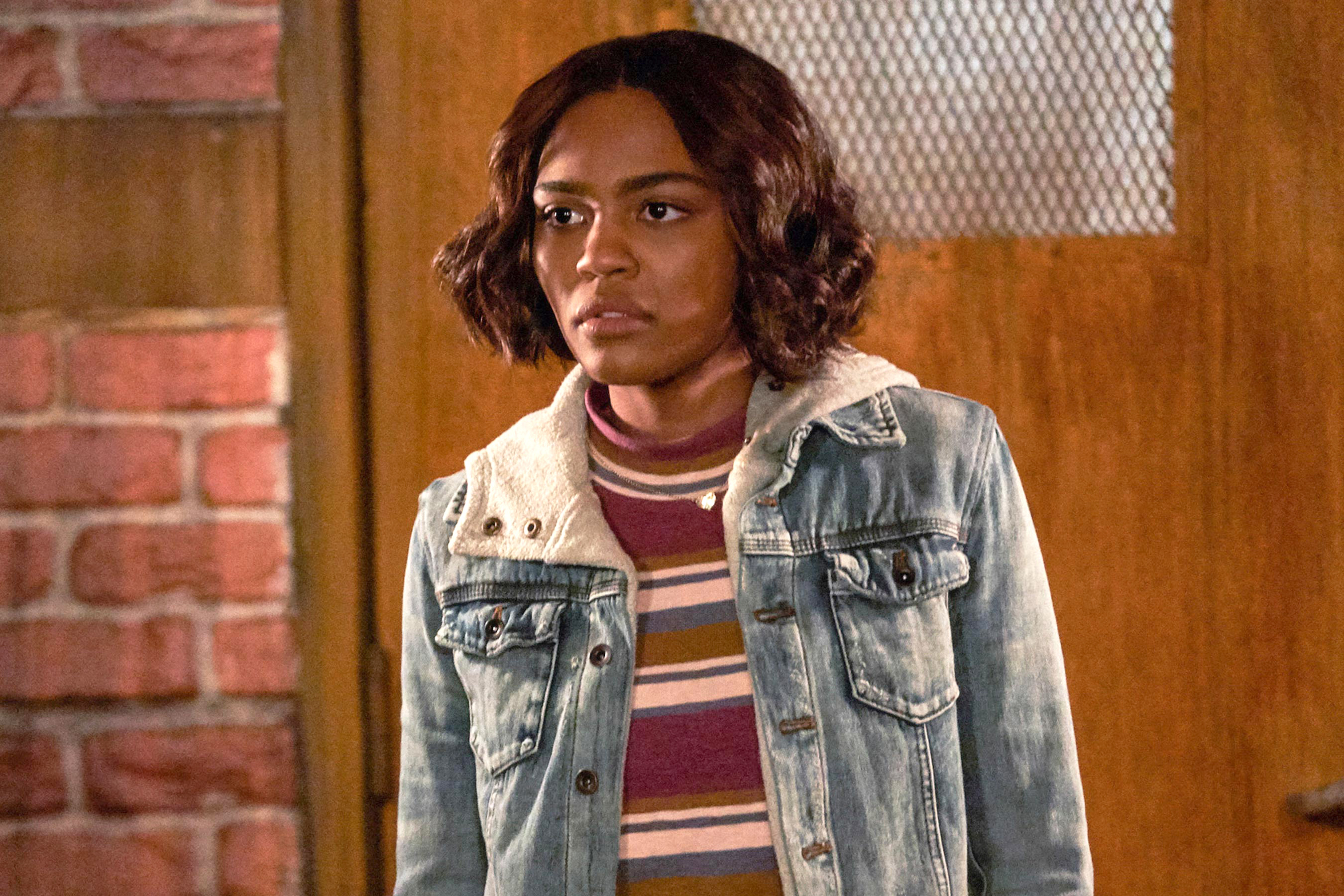 A girl (name unknown) stands in front of a door with a mesh window. She wears a striped shirt under a fur-lined denim jacket