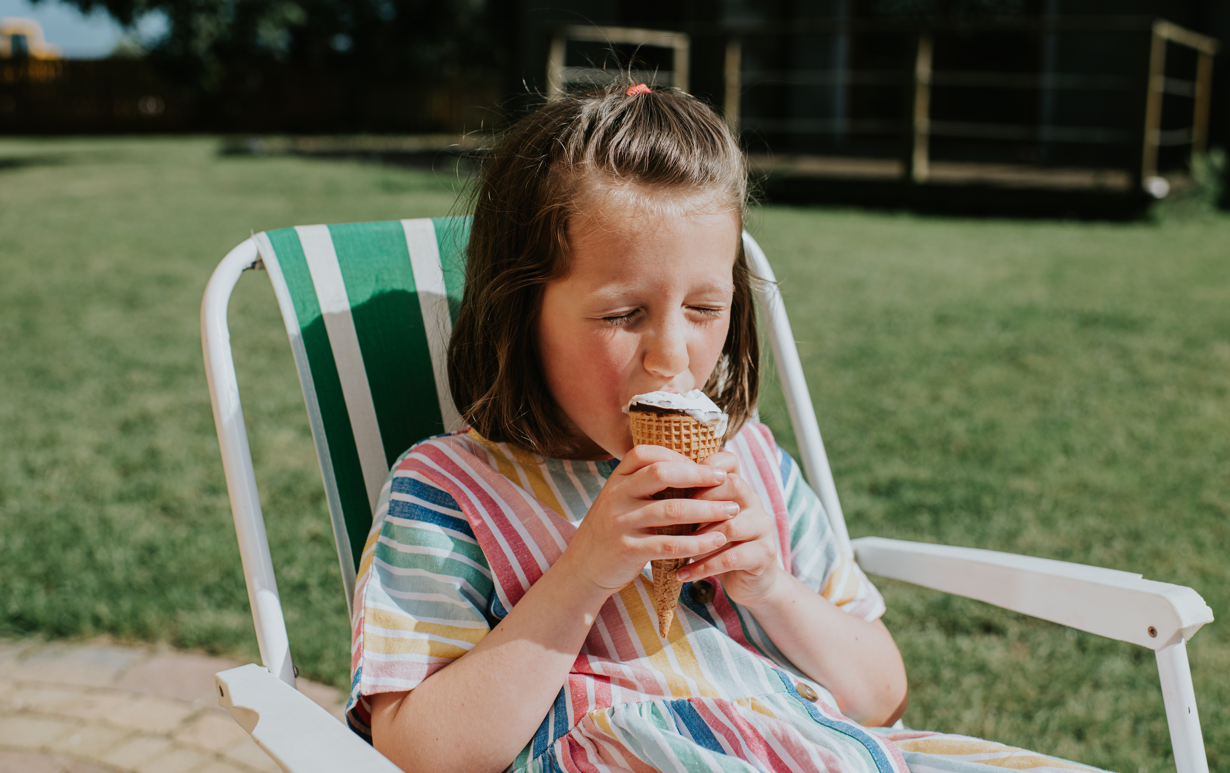 A young girl sits on a striped chair outdoors, enjoying an ice cream cone. She has short hair and wears a striped dress