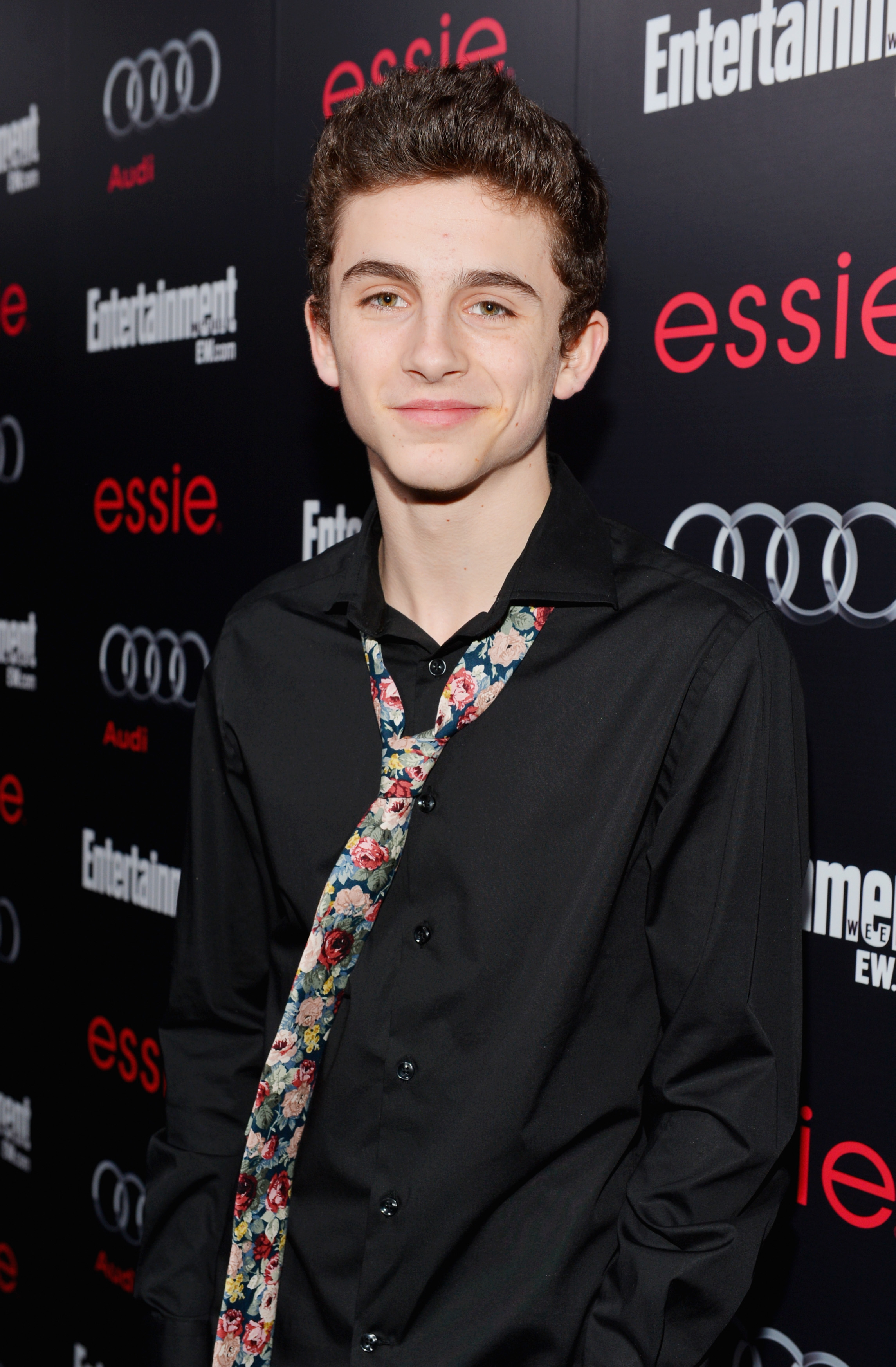 Timothée Chalamet on the red carpet in a black shirt with a loosely tied, floral-printed tie, attending an event featuring Audi, Entertainment Weekly, and essie branding