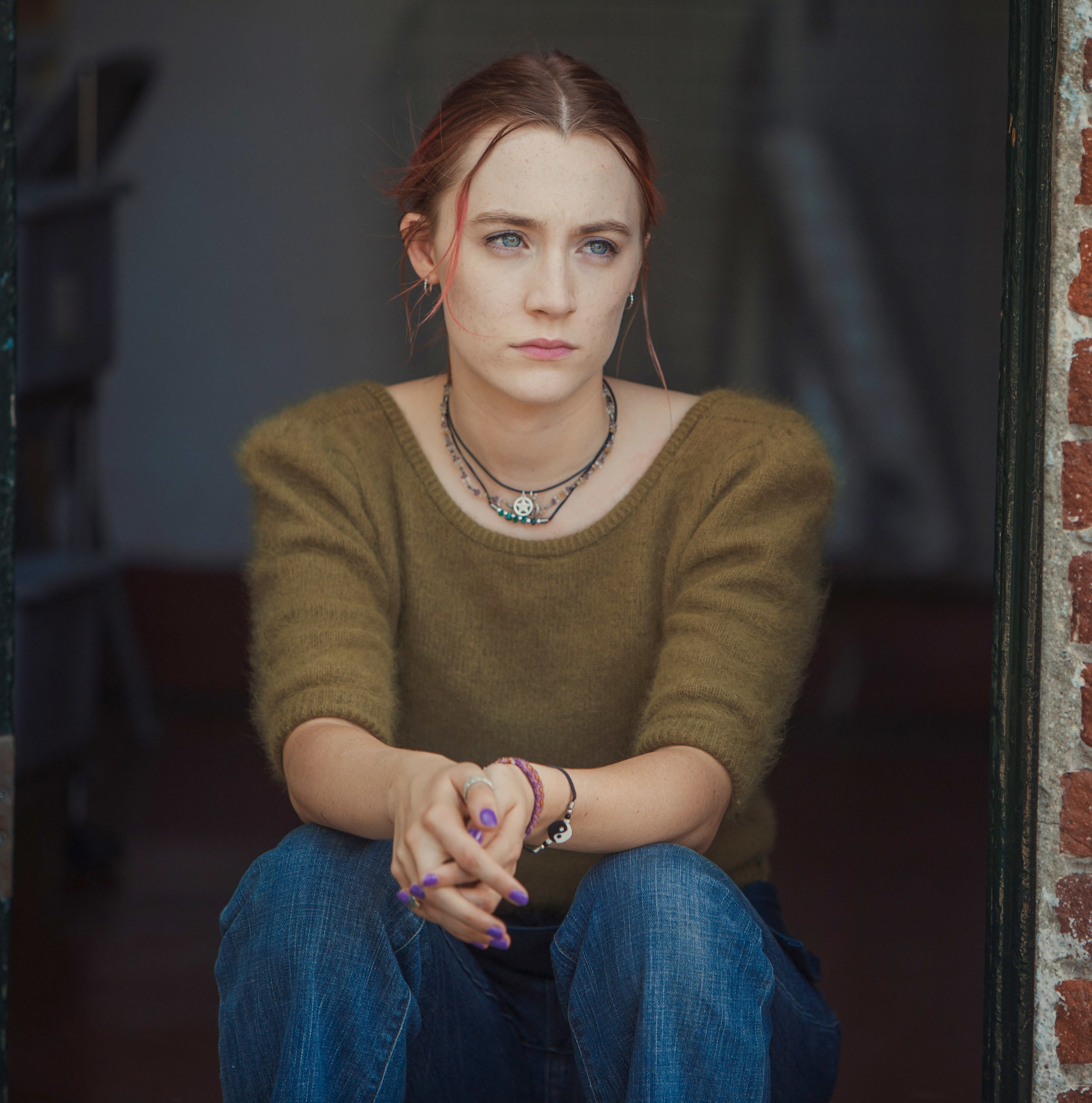 Saoirse Ronan sitting in a doorway, wearing a casual outfit with a green top and blue jeans, looking contemplative