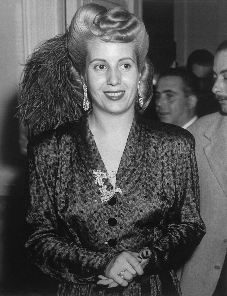 Woman in a patterned dress with ornate earrings and a feathered hairstyle, standing and smiling. Several people are blurred in the background