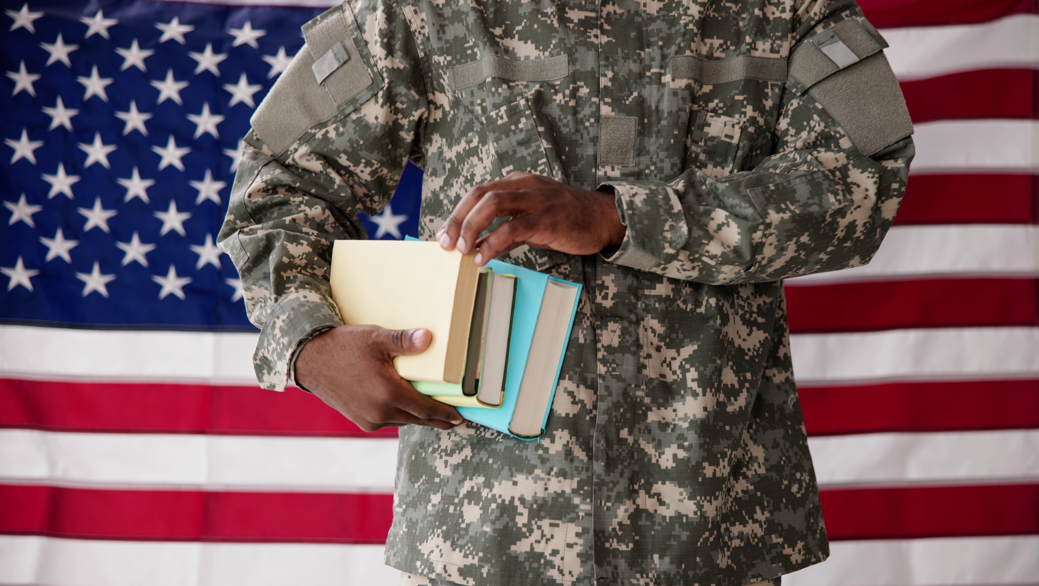 A person in a military uniform holds several books against a backdrop of the American flag