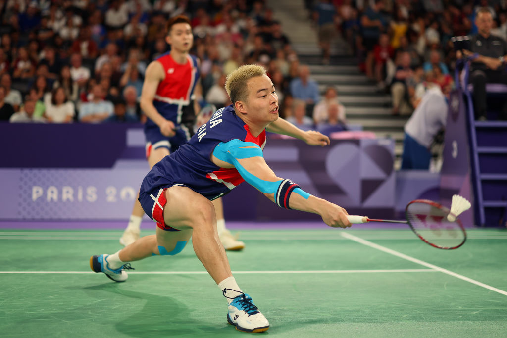 Badminton player Aaron Chia of Team Malaysia lunges to hit the shuttlecock during a competitive match at Paris 2024, with another player positioned behind him. Crowd watches intently
