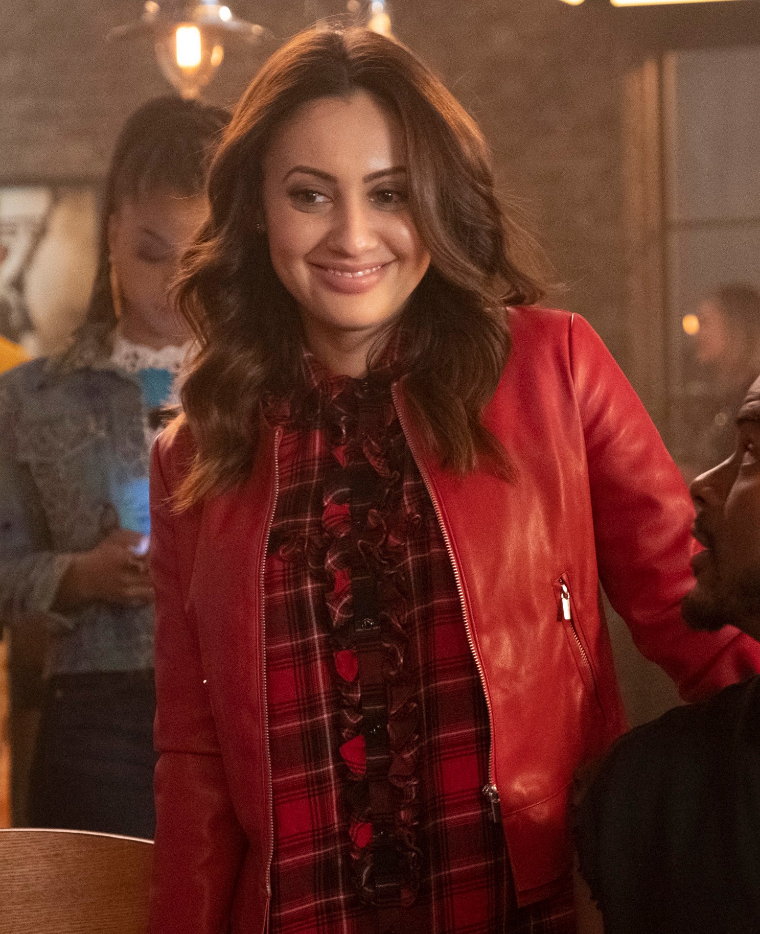 Francia Raisa stands smiling in a bar, wearing a red leather jacket and patterned dress, while looking at a seated Sarunas J. Jackson