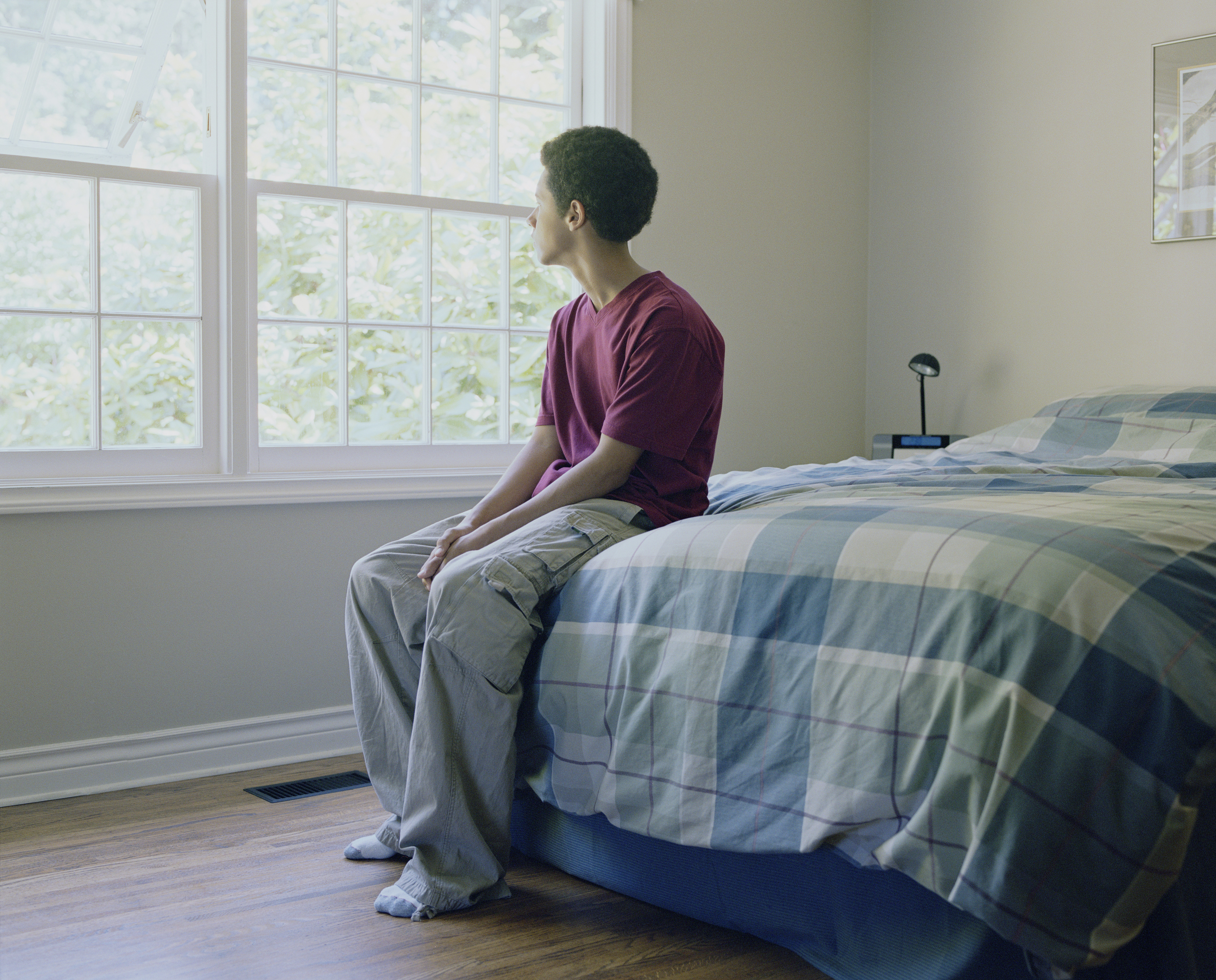 A teenager is sitting on a bed in a bedroom, looking out the window. The person is wearing casual clothing and appears deep in thought. The room is simply furnished