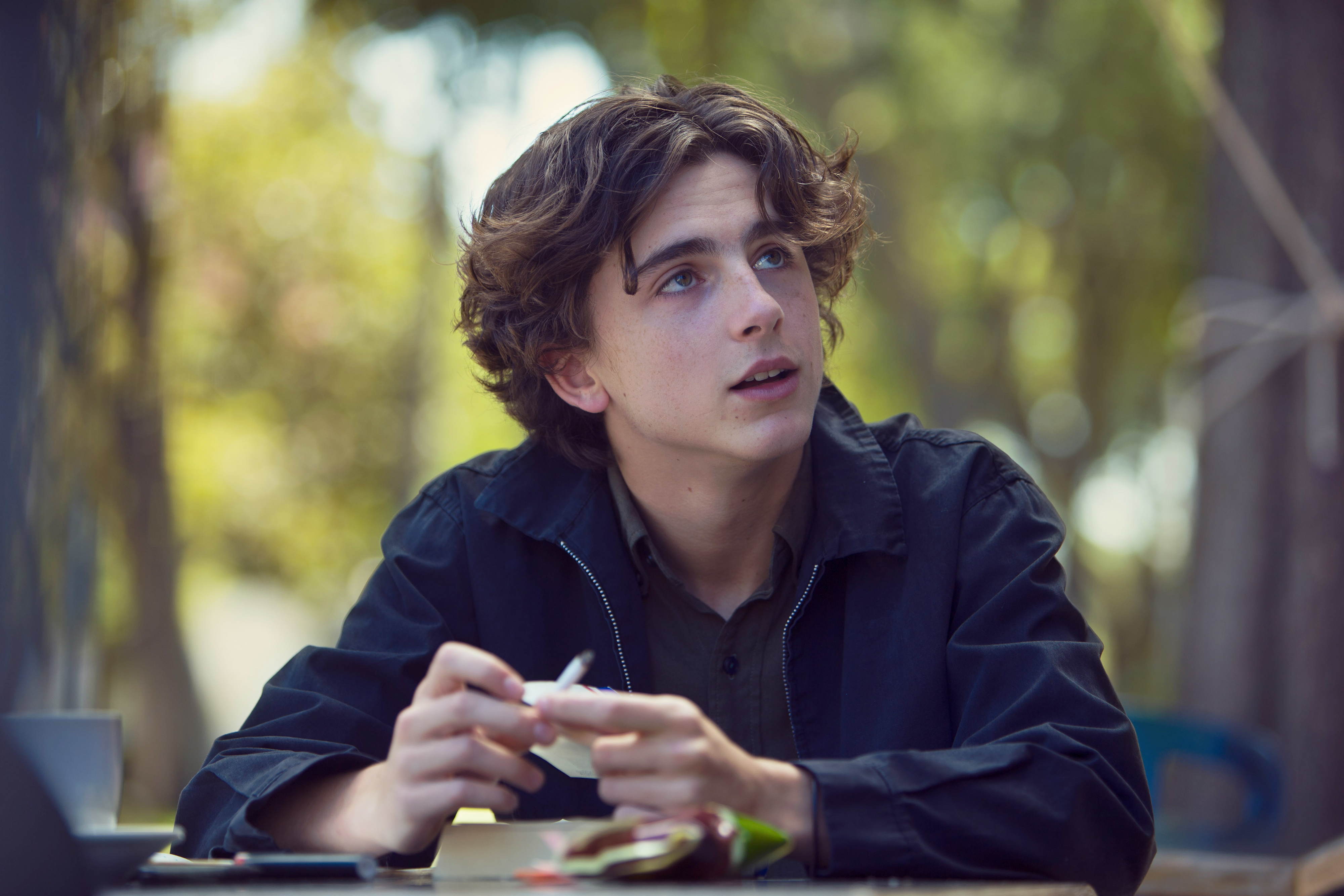 Timothée Chalamet is sitting outdoors at a table, holding an object and looking upwards. He is wearing a casual jacket