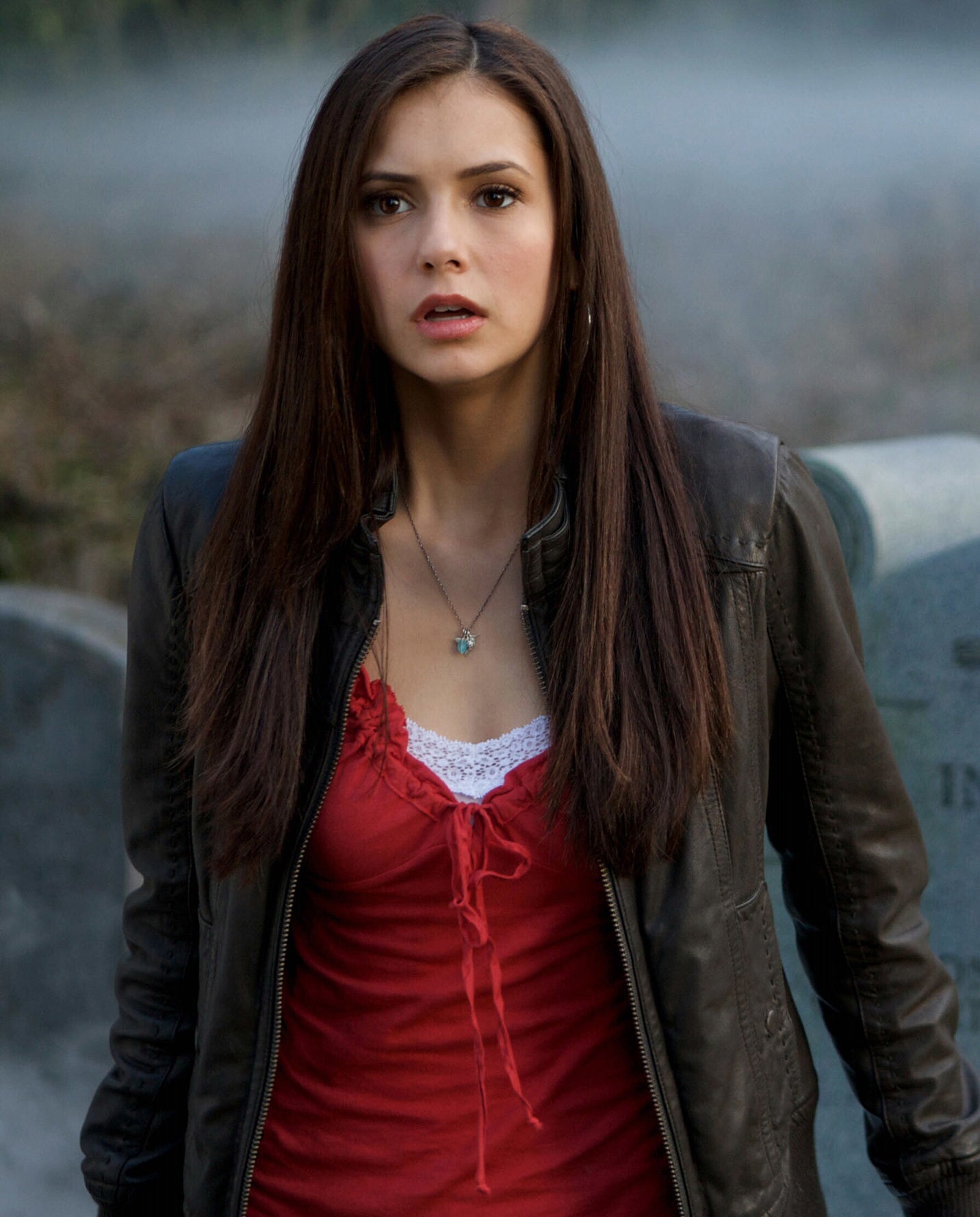 Nina Dobrev stands in front of a tombstone in a graveyard, looking surprised. She wears a jacket over a top