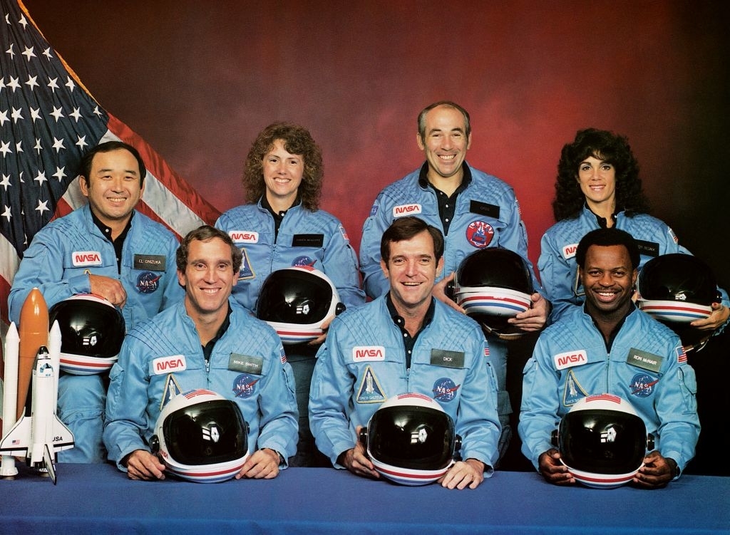 Group photo of seven Challenger astronauts wearing NASA spacesuits and holding helmets. Pictured are Ellison Onizuka, Christa McAuliffe, Gregory Jarvis, Judith Resnik, Michael Smith, Dick Scobee, and Ronald McNair