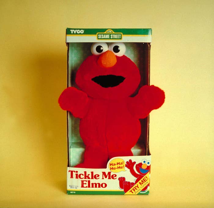 Tickle Me Elmo toy in its original packaging from Tyco, with Sesame Street branding and Try Me label. Elmo's hands are raised and the box features "Ha-Ha! He-He!" text