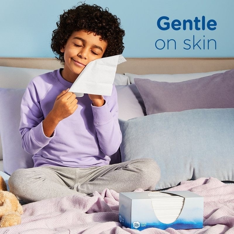 Child sits on a bed, smiling with eyes closed while holding a tissue to their face. Text reads "Gentle on skin." A tissue box is on the bed