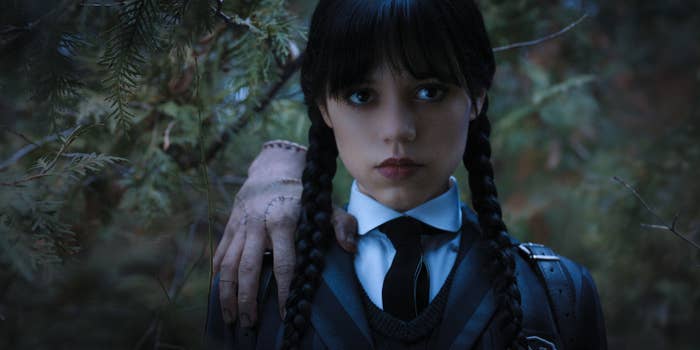 Jenna Ortega as Wednesday Addams in a still from the show "Wednesday," wearing a school uniform with braided hair and a disembodied hand on her shoulder