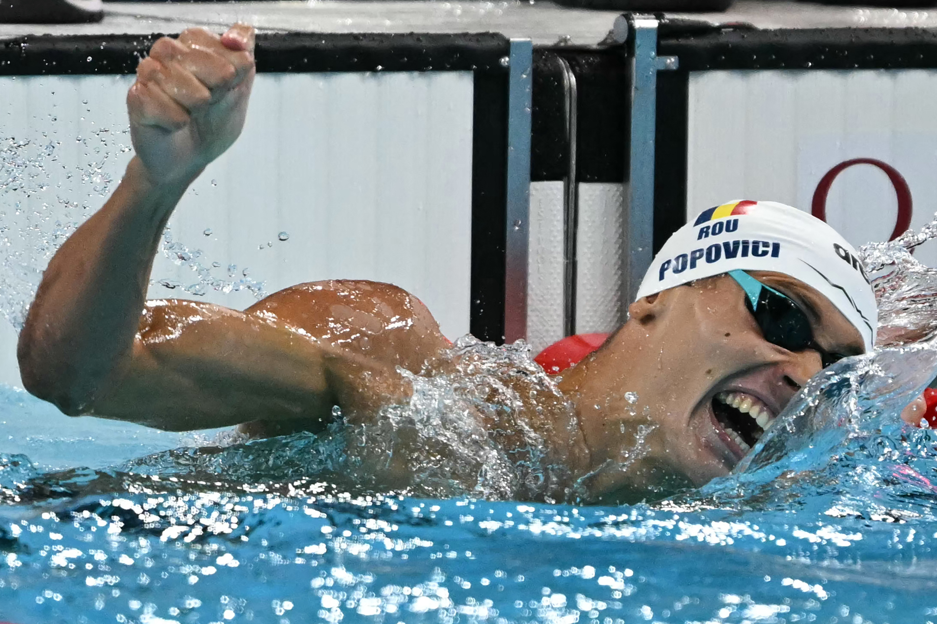 David Popovici celebrates in a pool, wearing swim goggles and a cap with &quot;ROU&quot; and &quot;Popovici&quot; on it