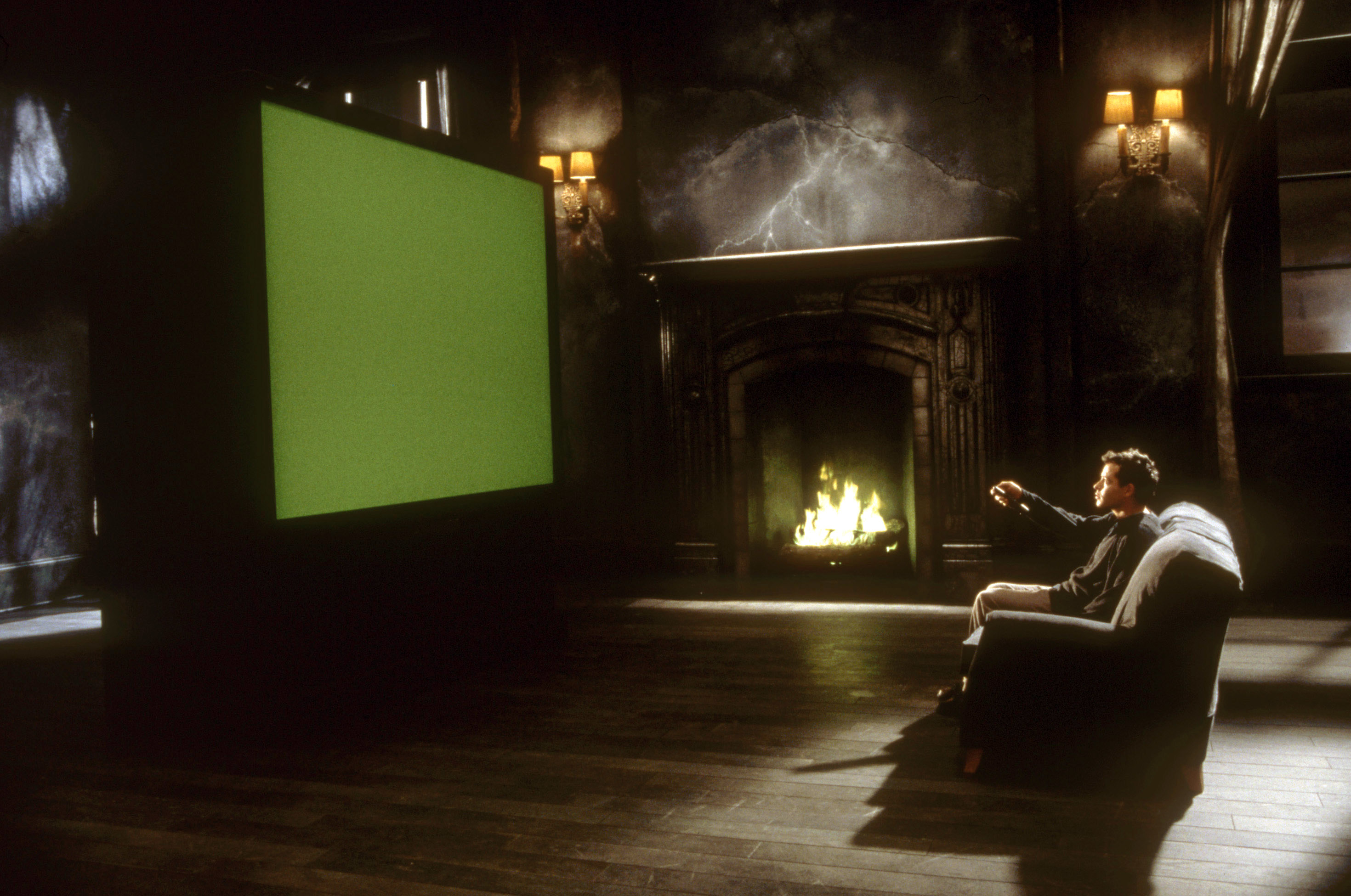 A person sits in a dark room on a couch, pointing at a screen on the left. A lit fireplace is in the background
