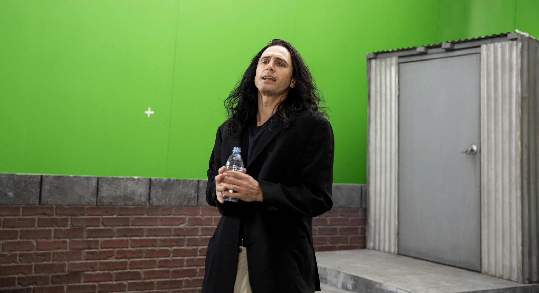James Franco stands in front of a green screen and brick wall, holding a water bottle. He is dressed in a black jacket and beige pants