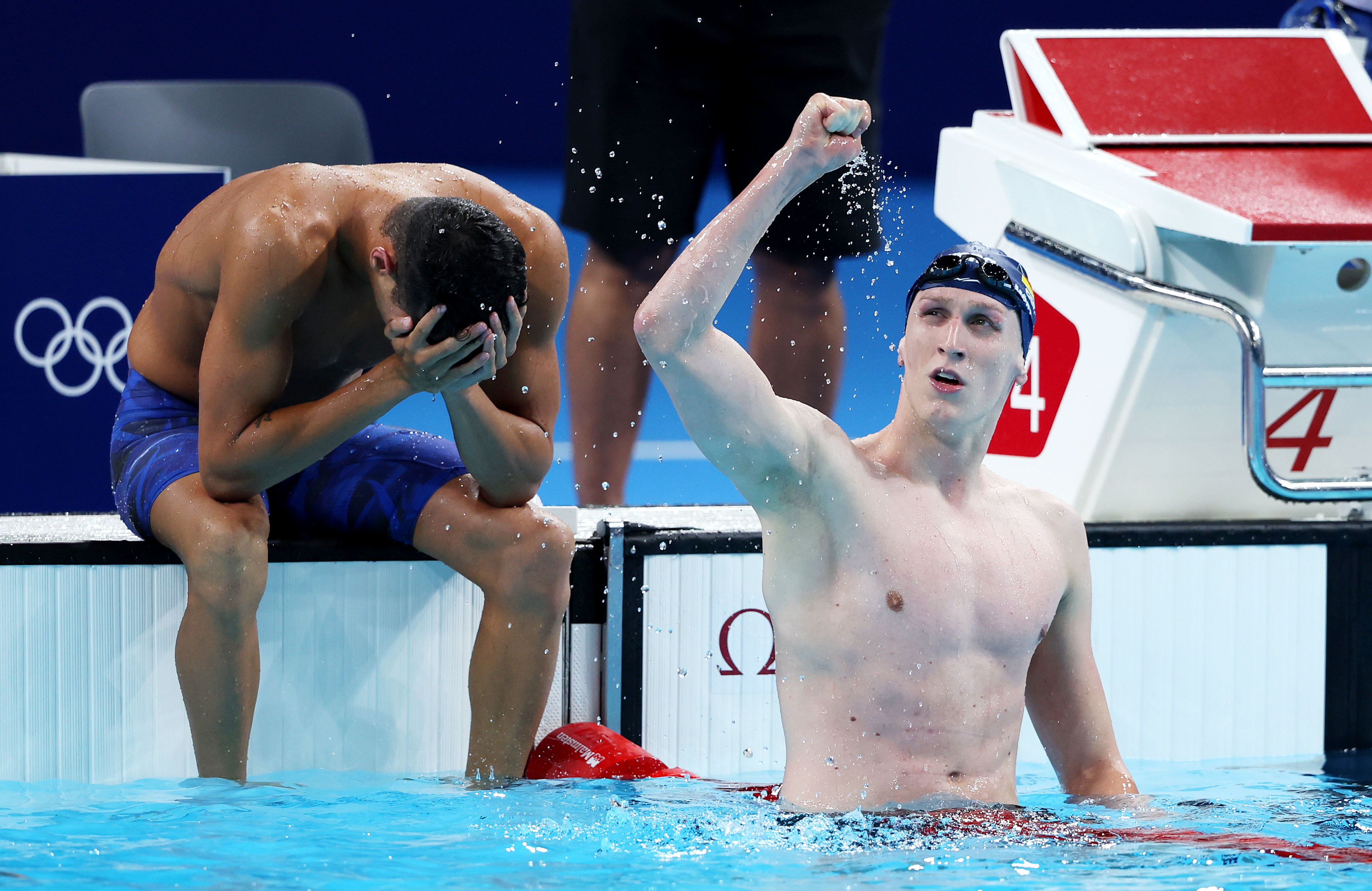 Two swimmers at the Olympics; one is seated with his head in his hands, while the other raises his fist in celebration