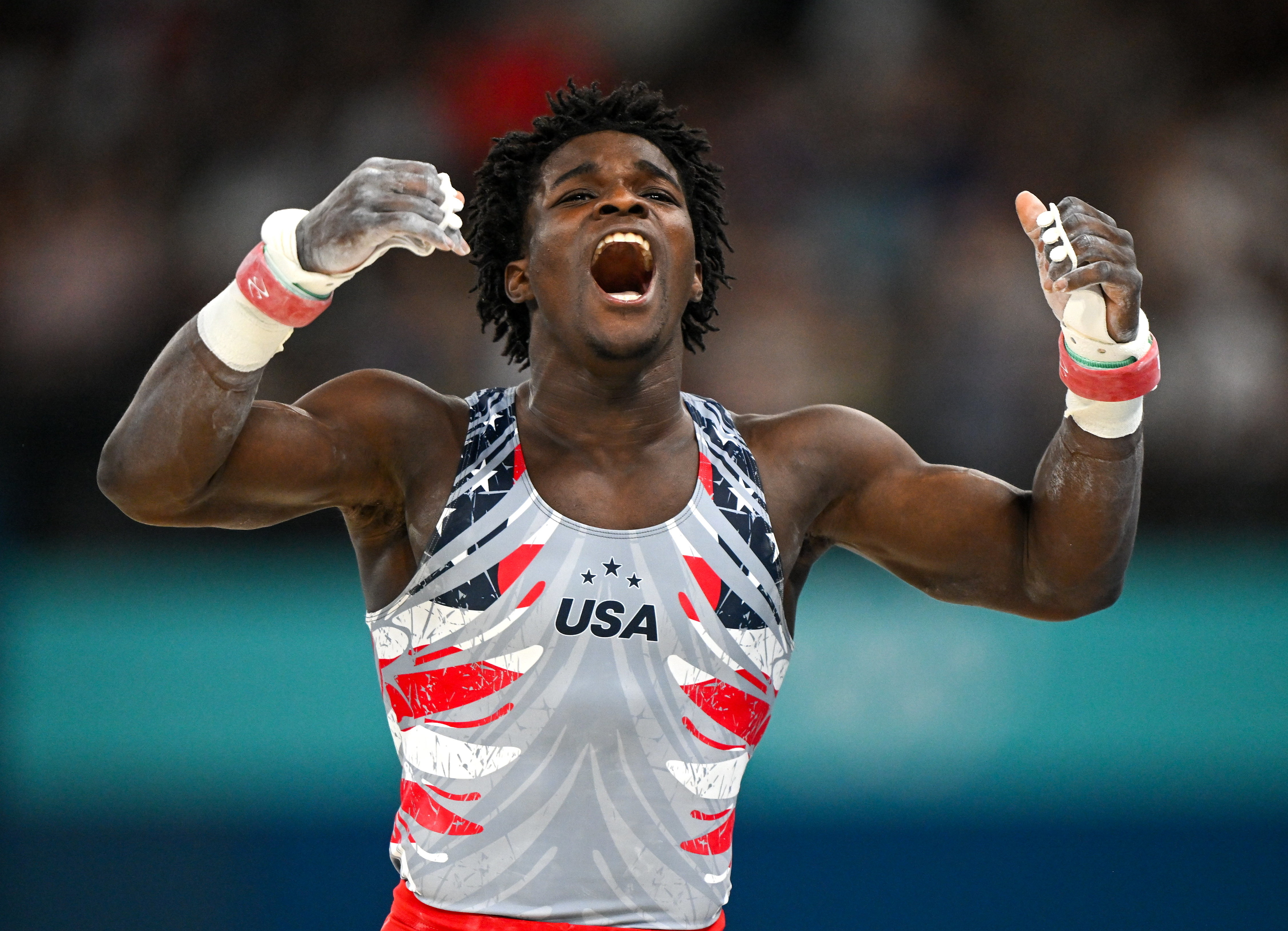 An athlete wearing a 'USA' tank top celebrates with arms raised and mouth open in triumph at a sports event