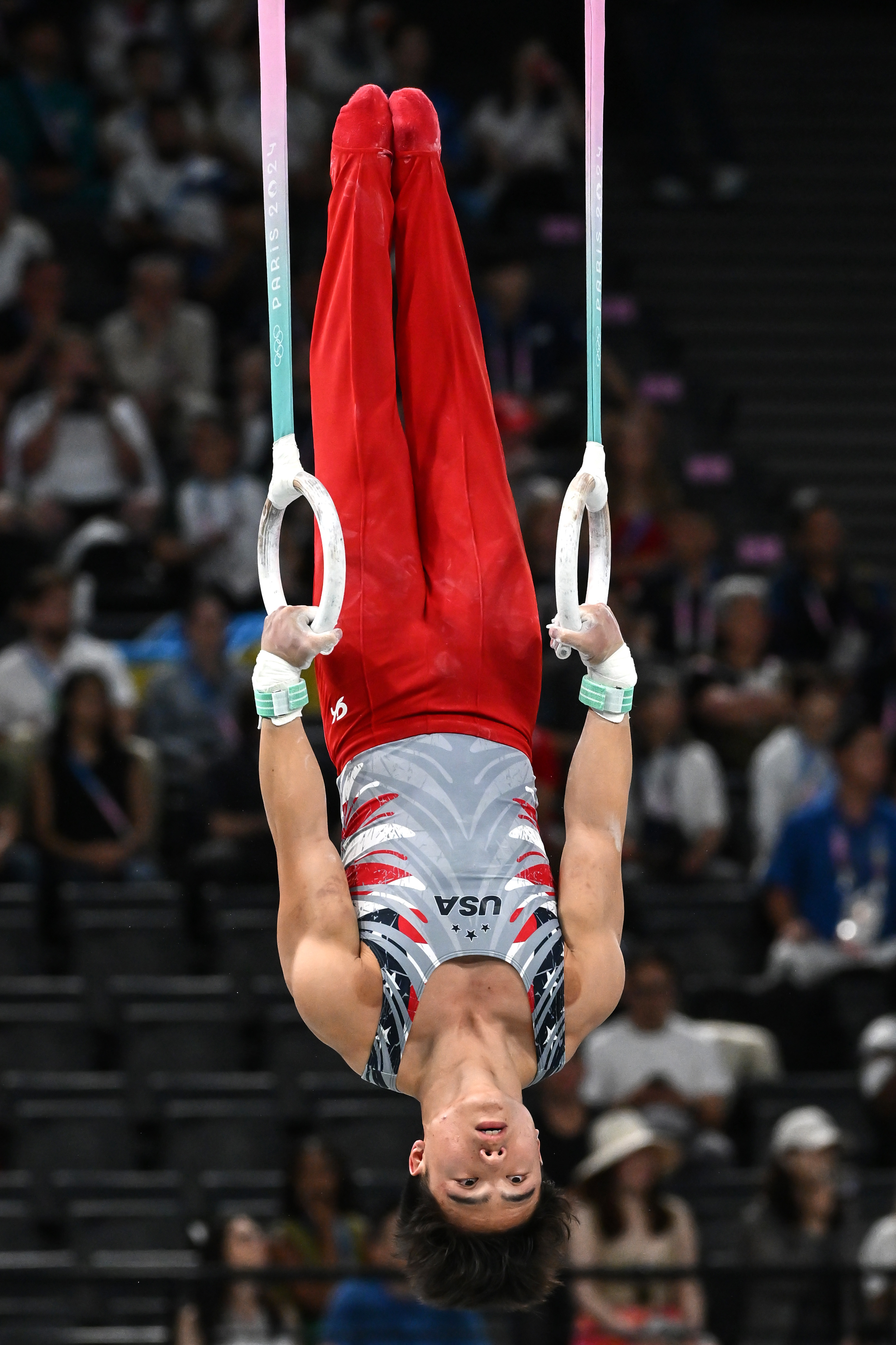 Kōhei Uchimura performs a gymnastics routine on the rings, wearing a form-fitting USA-branded leotard with red pants