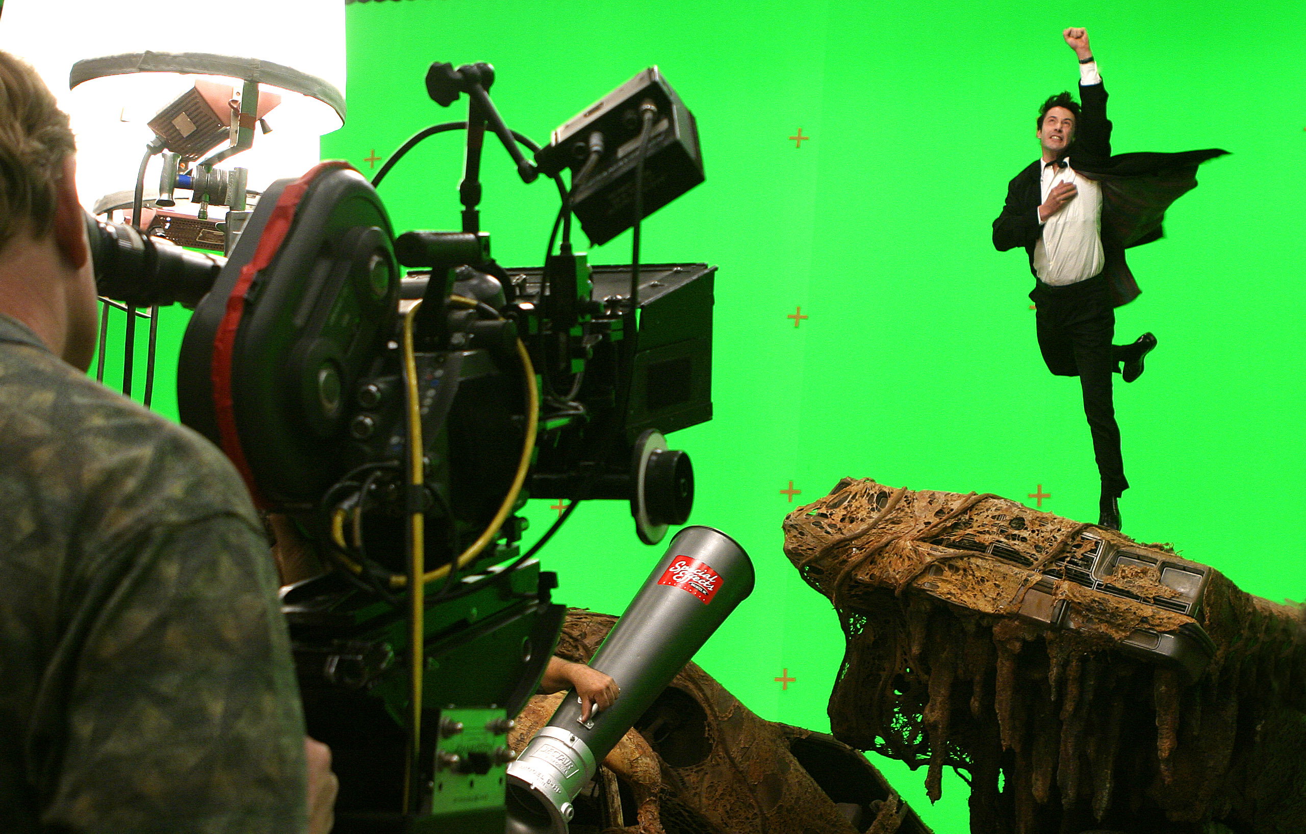 Actor in a black suit performs an action scene against a green screen background, while film crew members operate a camera and lighting equipment on set