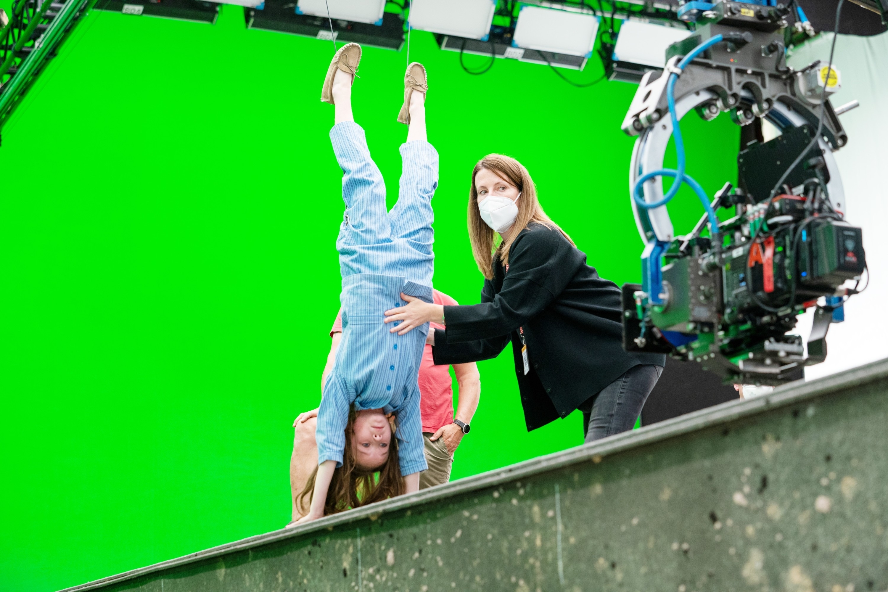 A young girl wearing casual clothing is doing a handstand, supported by a masked woman, on a set with a green screen background
