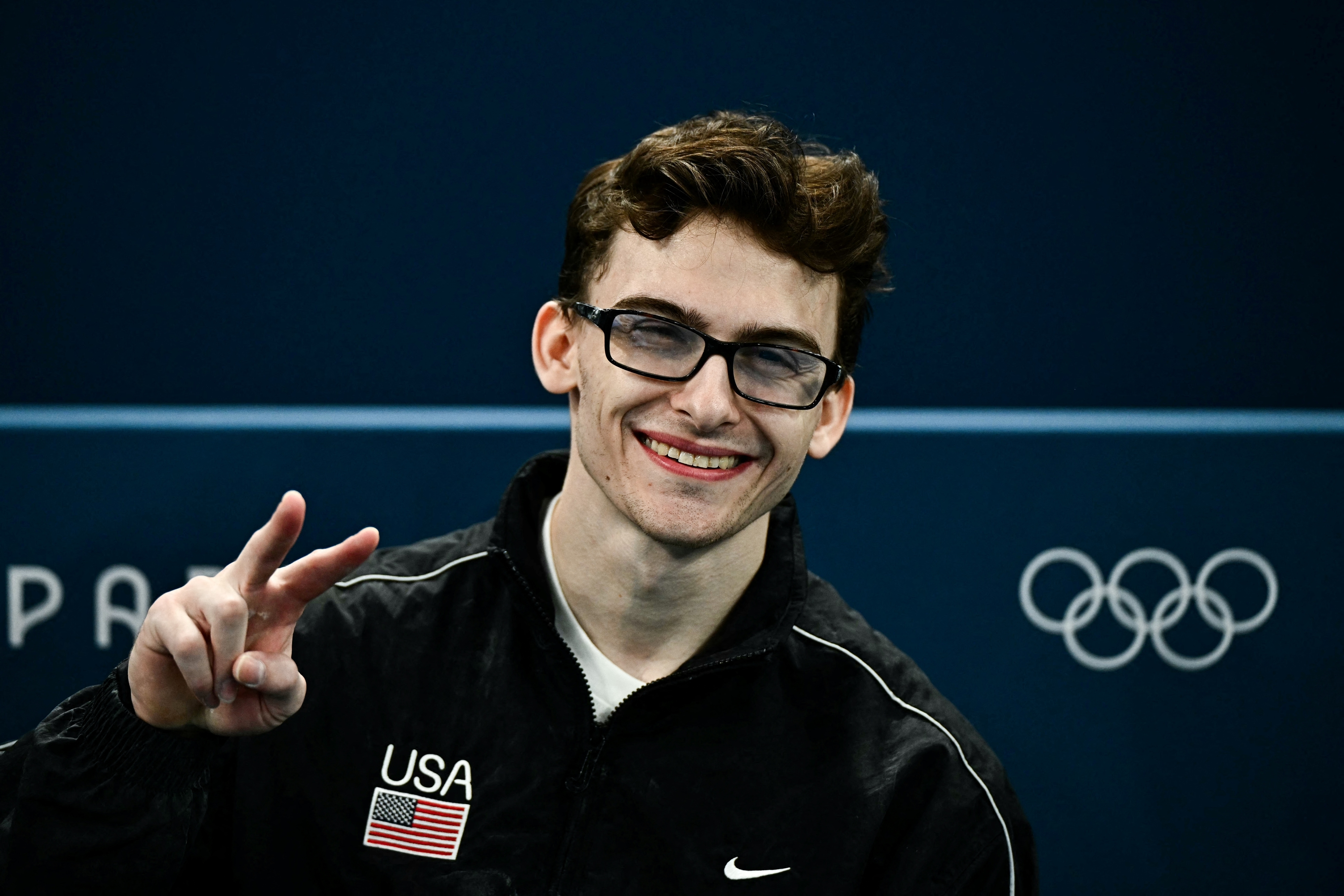 A man wearing a black USA jacket with an American flag patch smiles and makes a peace sign. Olympic rings are visible in the background. Names are not known