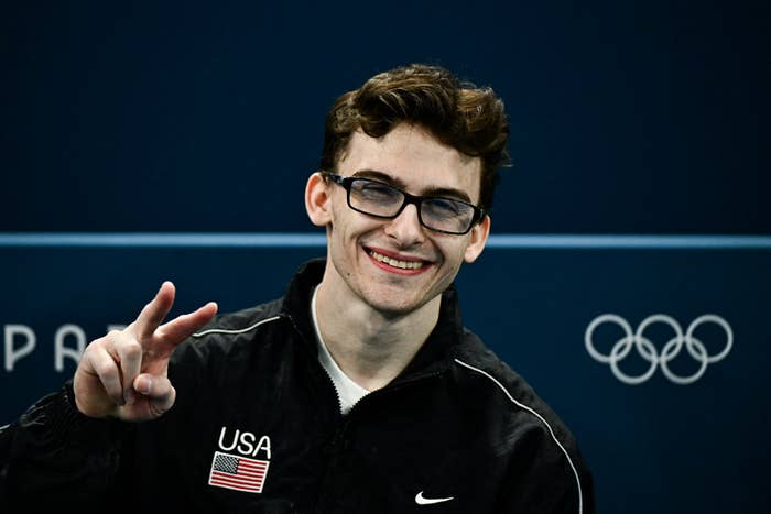 A antheral   wearing a achromatic  USA overgarment   with an American emblem  spot  smiles and makes a bid    sign. Olympic rings are disposable   successful  the background. Names are not known