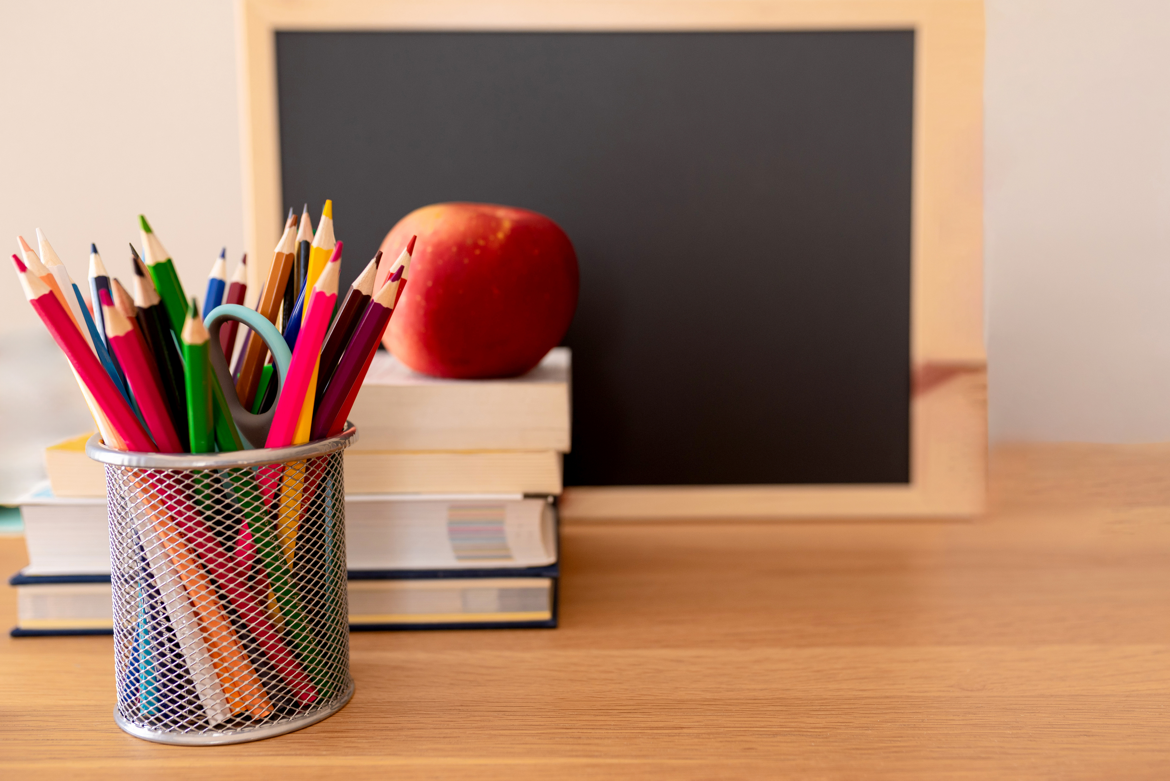 A pencil holder with colorful pencils, an apple, a stack of books, and a blank blackboard on a wooden desk