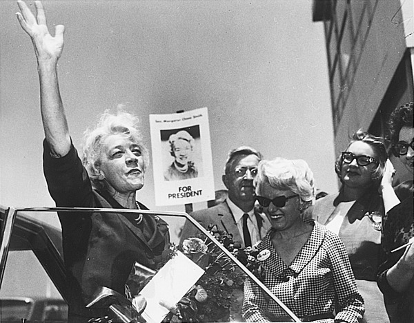 Margaret Chase Smith waves to the crowd while exiting a car, accompanied by supporters. A campaign poster behind her reads "Margaret Chase Smith For President."
