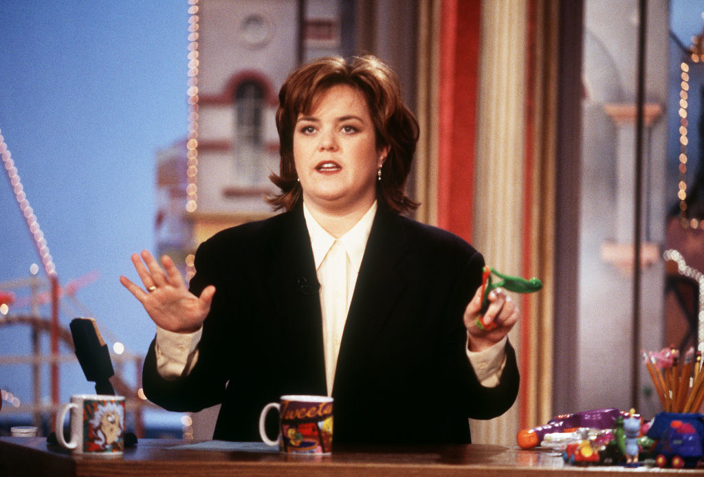 Rosie O'Donnell wears a black blazer and white shirt, holding a toy while hosting her talk show in the 1990s