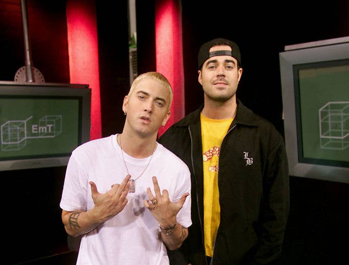 Eminem and Paul Rosenberg stand in a studio. Eminem poses with hand gestures, wearing a white T-shirt. Paul wears a black jacket and a cap