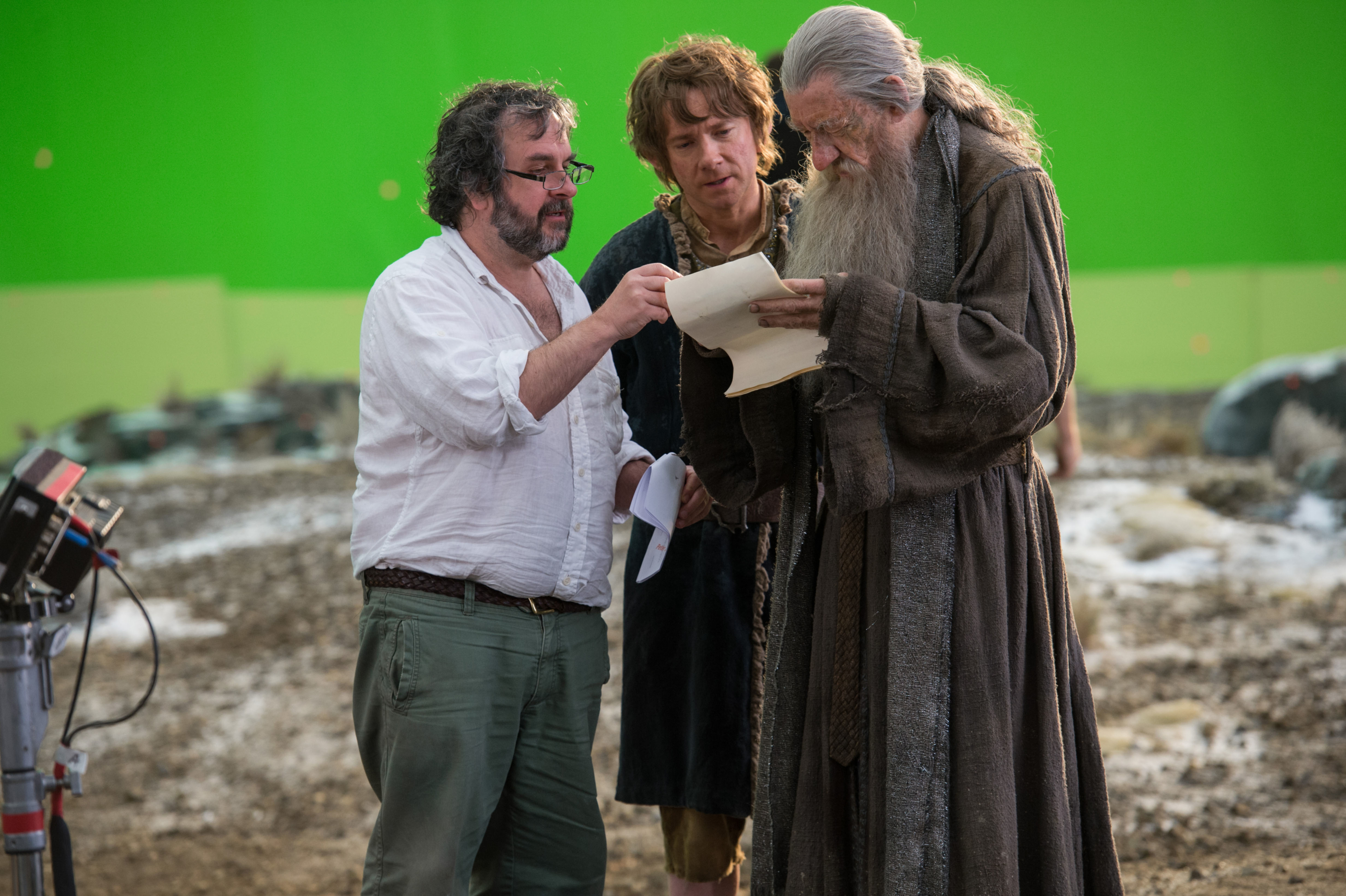 Peter Jackson, Elijah Wood, and Ian McKellen review a script on a movie set. Ian McKellen is in costume as Gandalf from "The Lord of the Rings."