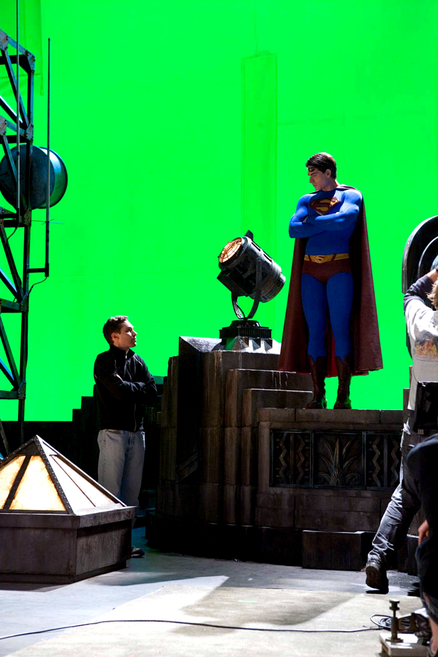 Man in casual clothes talks to a person dressed as Superman on a movie set with a green screen background