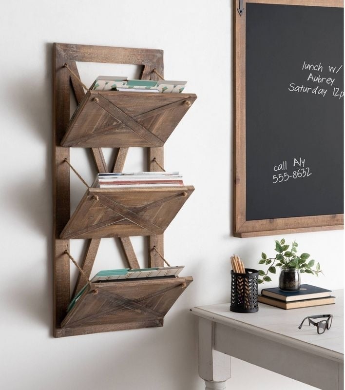 Wooden wall-mounted magazine rack filled with books and magazines, next to a white desk with decor, and a chalkboard with a note to call Aly at 555-8632