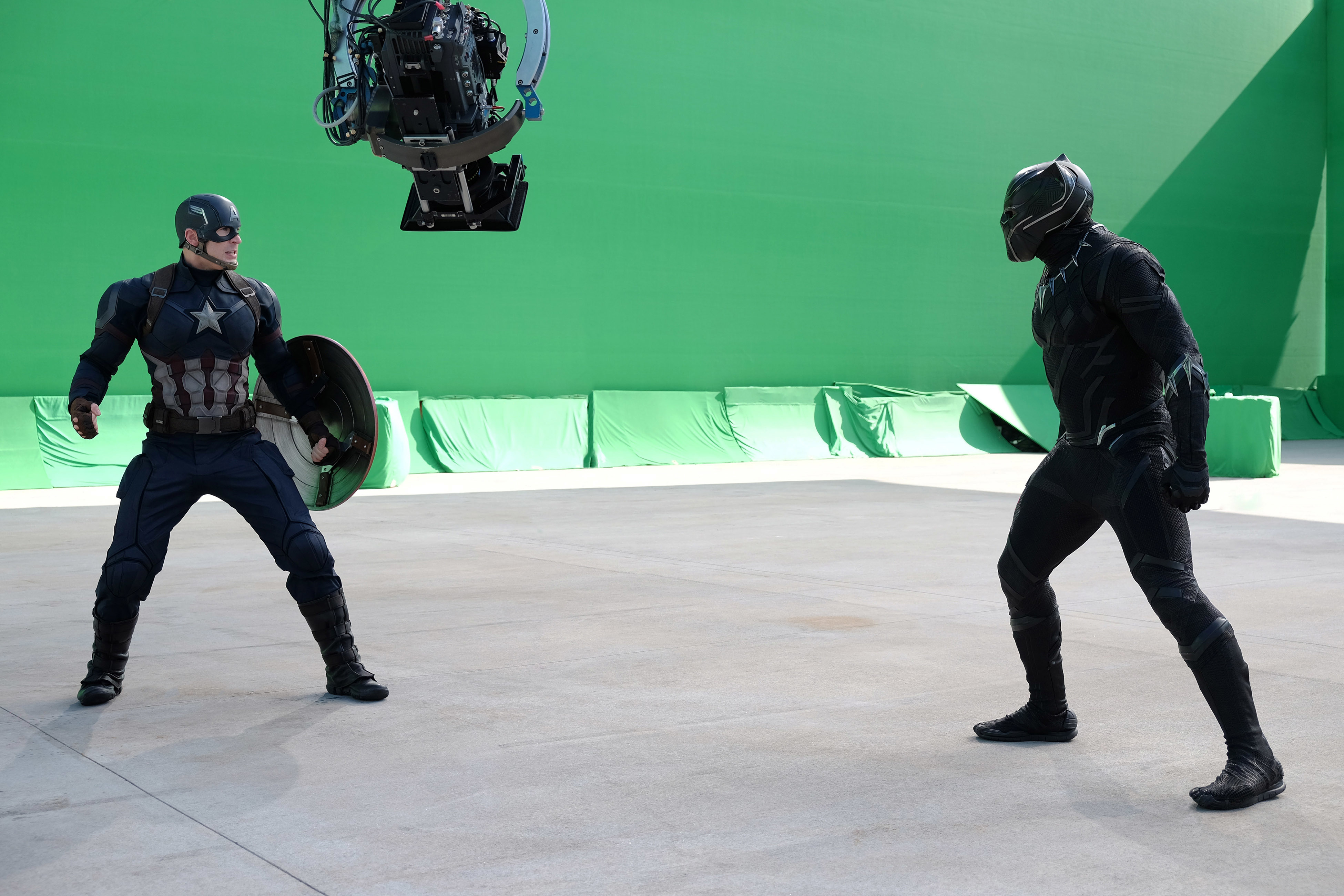 Captain America and Black Panther face each other in costume on a film set with a green screen and camera rigging above