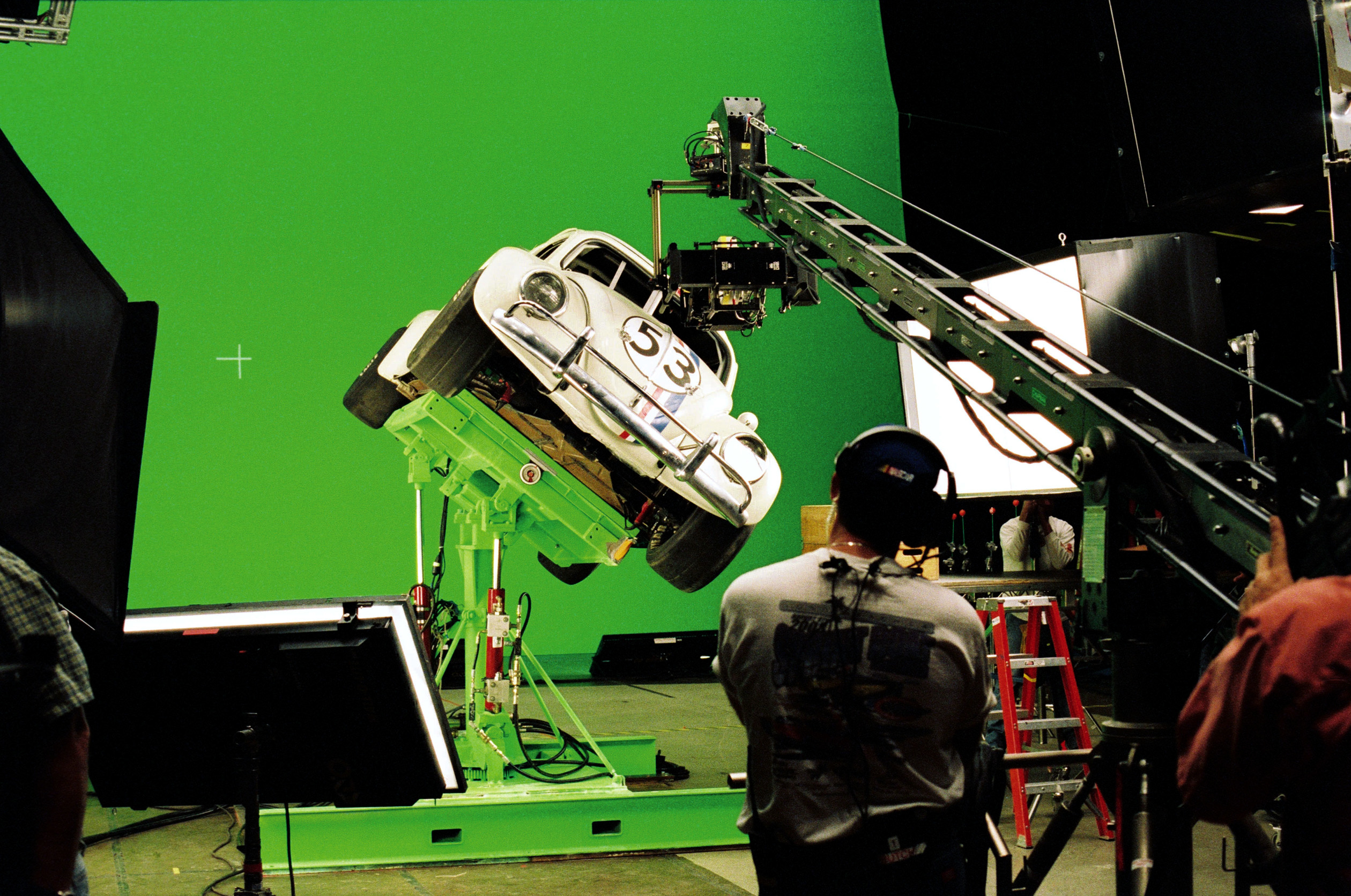 People working on a film set with a car mounted on a tilting platform against a green screen, indicating a behind-the-scenes look at special effects in production
