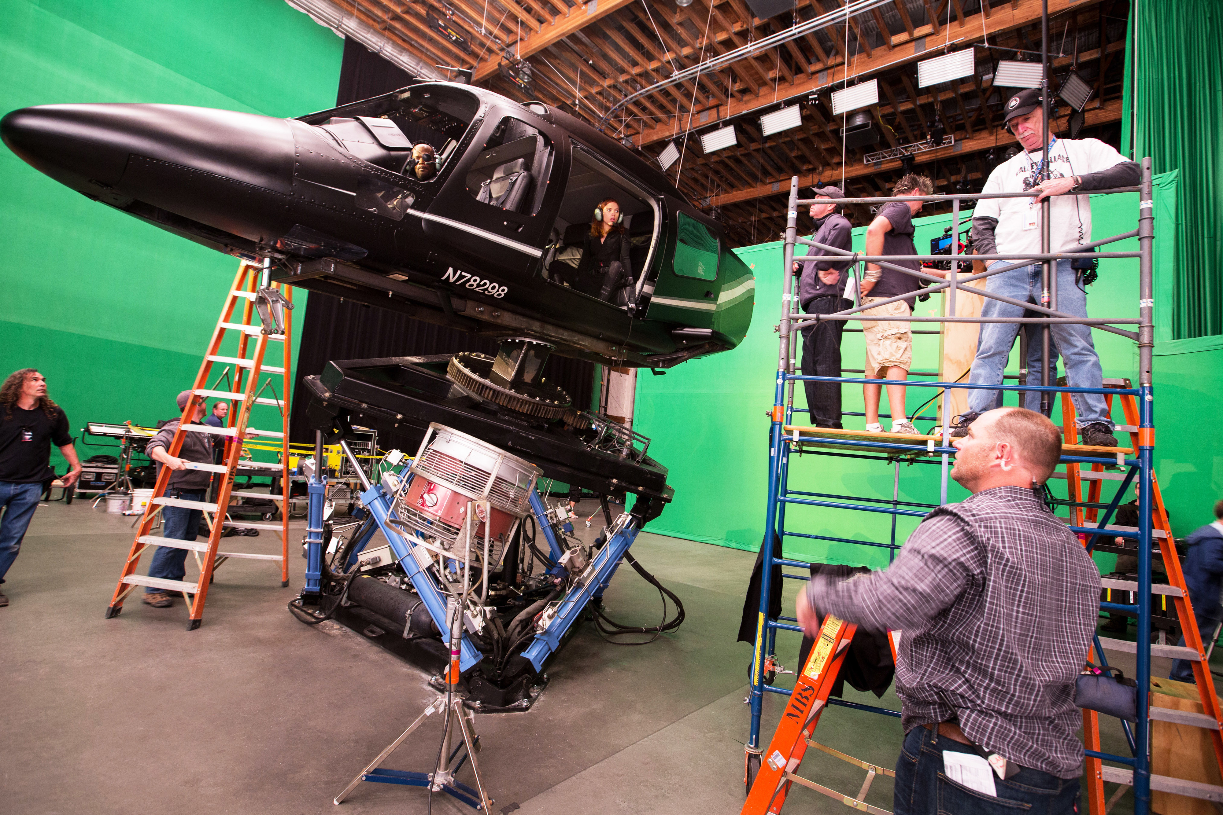 Film crew on set with a helicopter prop suspended by mechanical supports, surrounded by green screens, ladders, and scaffolding