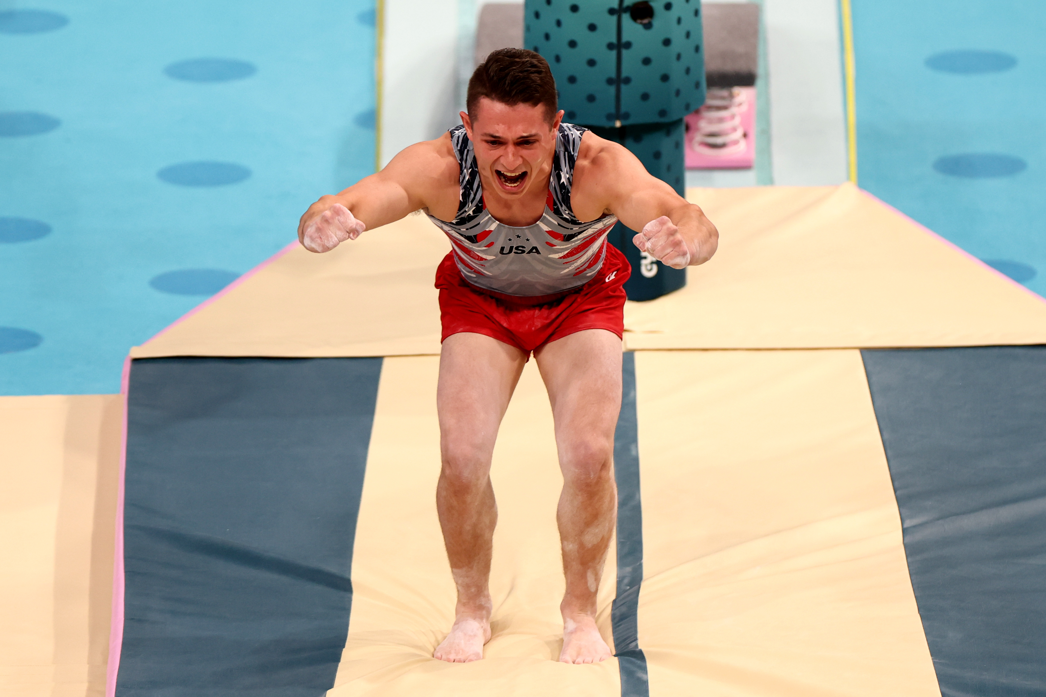A male gymnast wearing a USA uniform consisting of a sleeveless, patterned top and red shorts lands a routine with a focused expression in a gymnastics arena