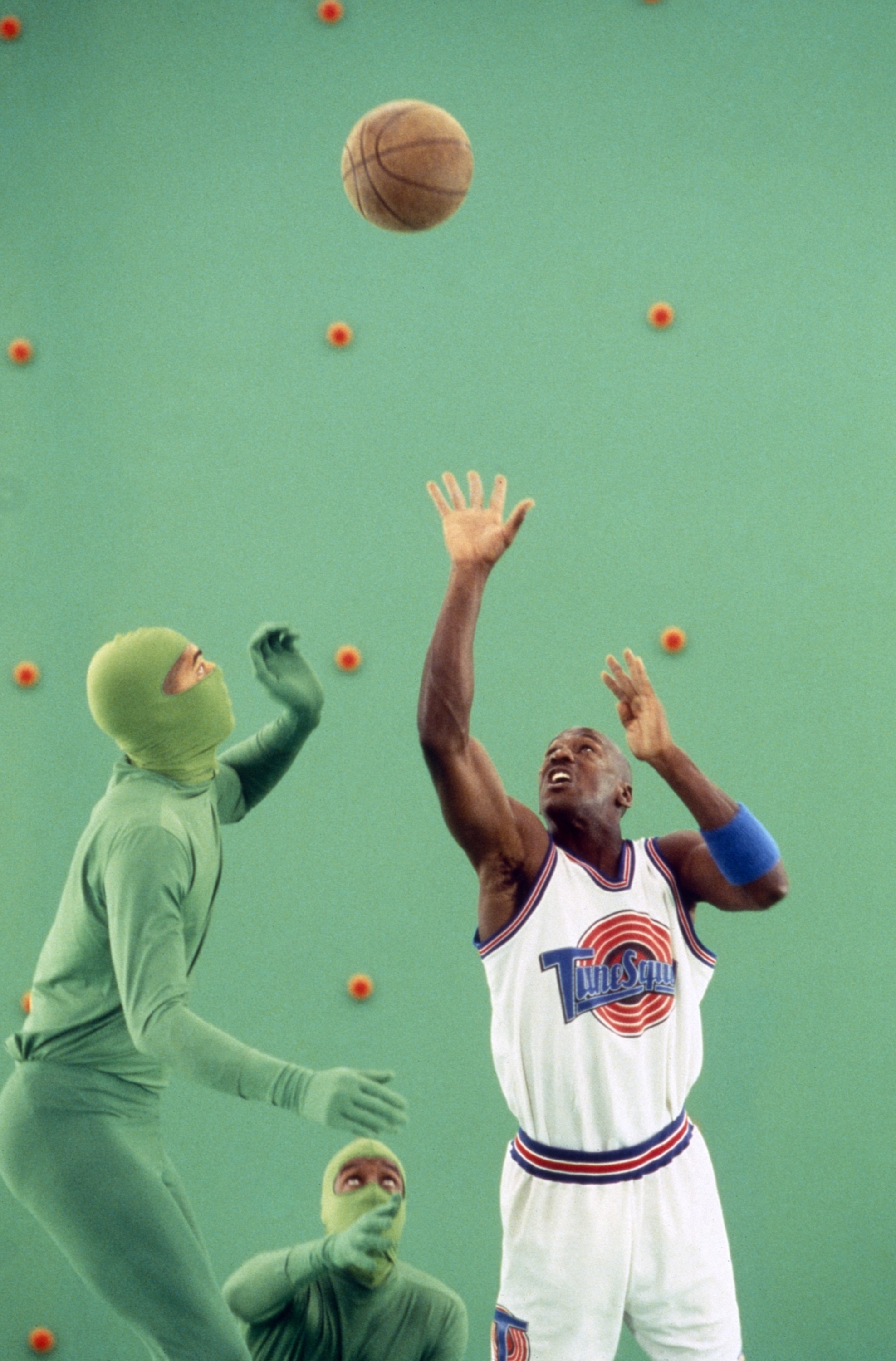 Michael Jordan, in a Tune Squad jersey, jumps to catch a basketball mid-air while surrounded by people in green suits during the filming of Space Jam