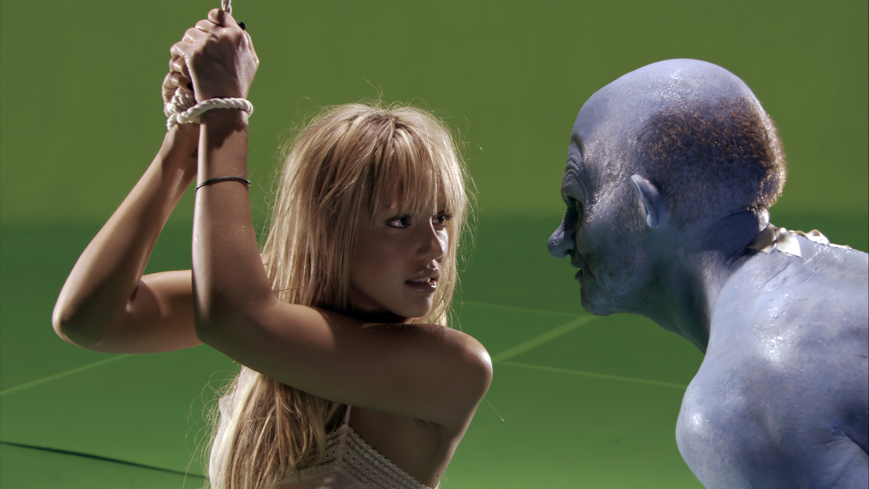 Jessica Alba, with her hands tied above her head, faces a person painted blue with prosthetic features in this movie scene