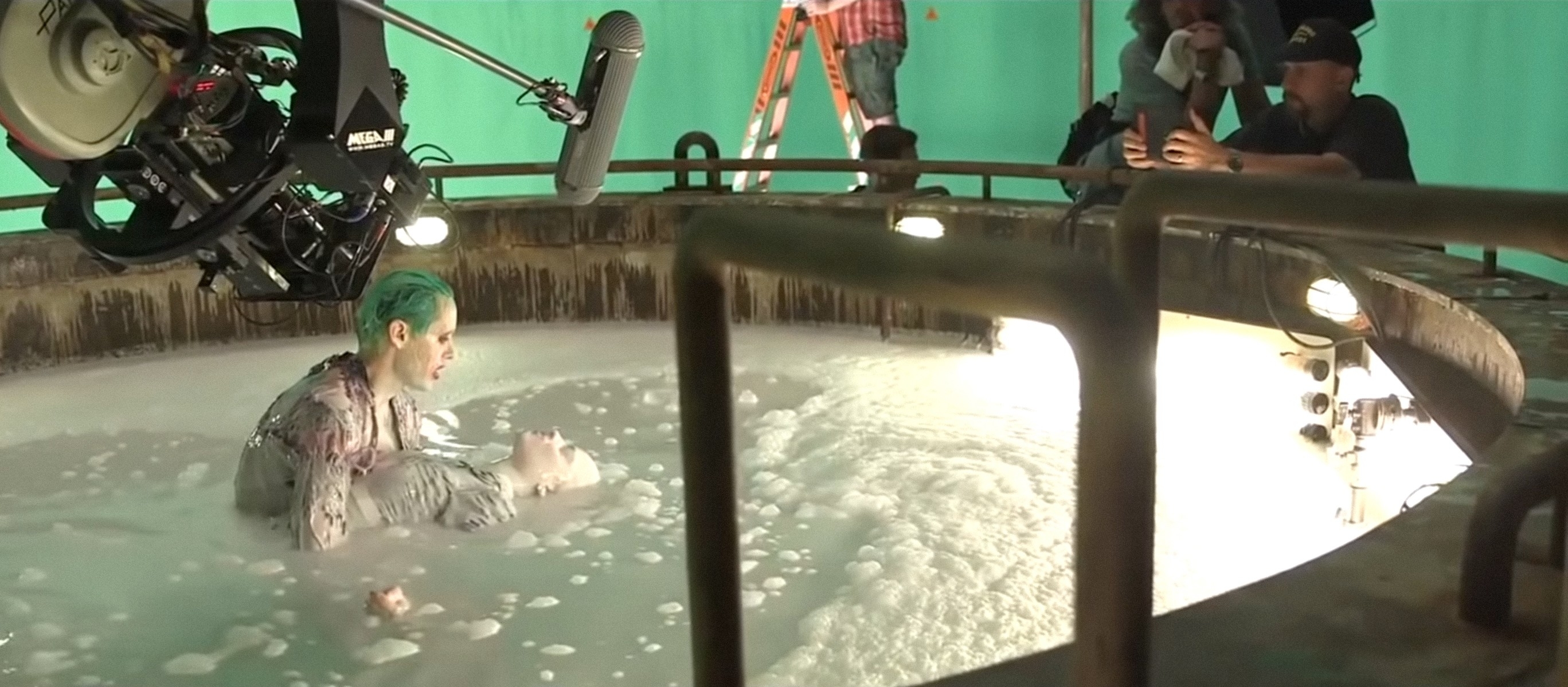 Jared Leto with green hair in a foam-filled tank on set, likely filming a scene as the Joker. Crew members are seen operating equipment in the background