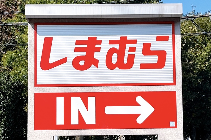 Sign with the text “しまむら IN →” indicating an entrance to Shimamura, a Japanese retail store, with trees and a street in the background