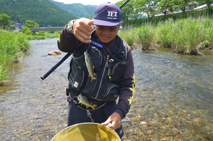 帽子と釣り用ベストを着た男性が、渓流で魚を釣り上げている。背景には草と木々が広がる。
