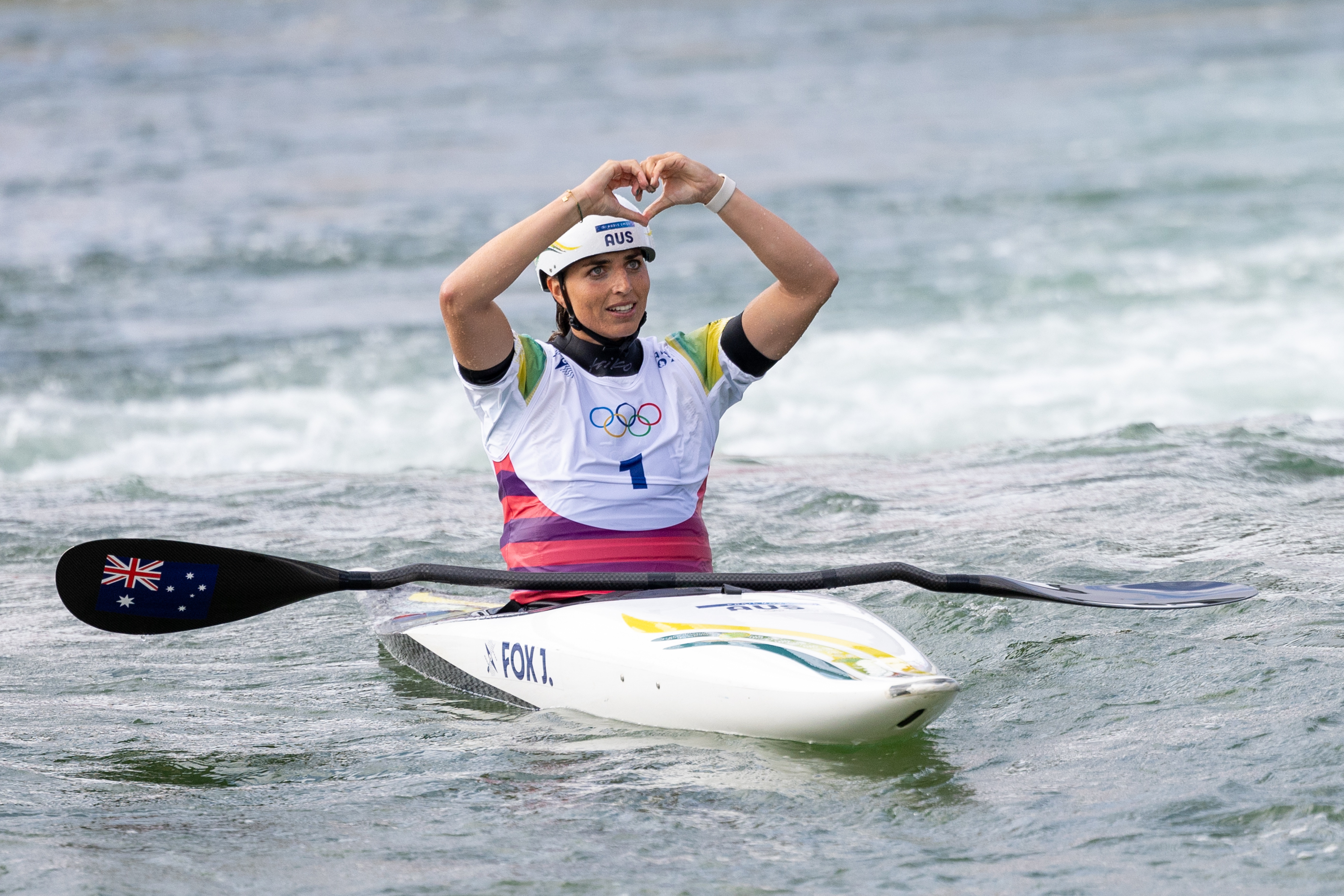 Jessica Fox celebrates in her kayak, making a heart shape with her hands after a race. She wears an Olympic jersey and helmet
