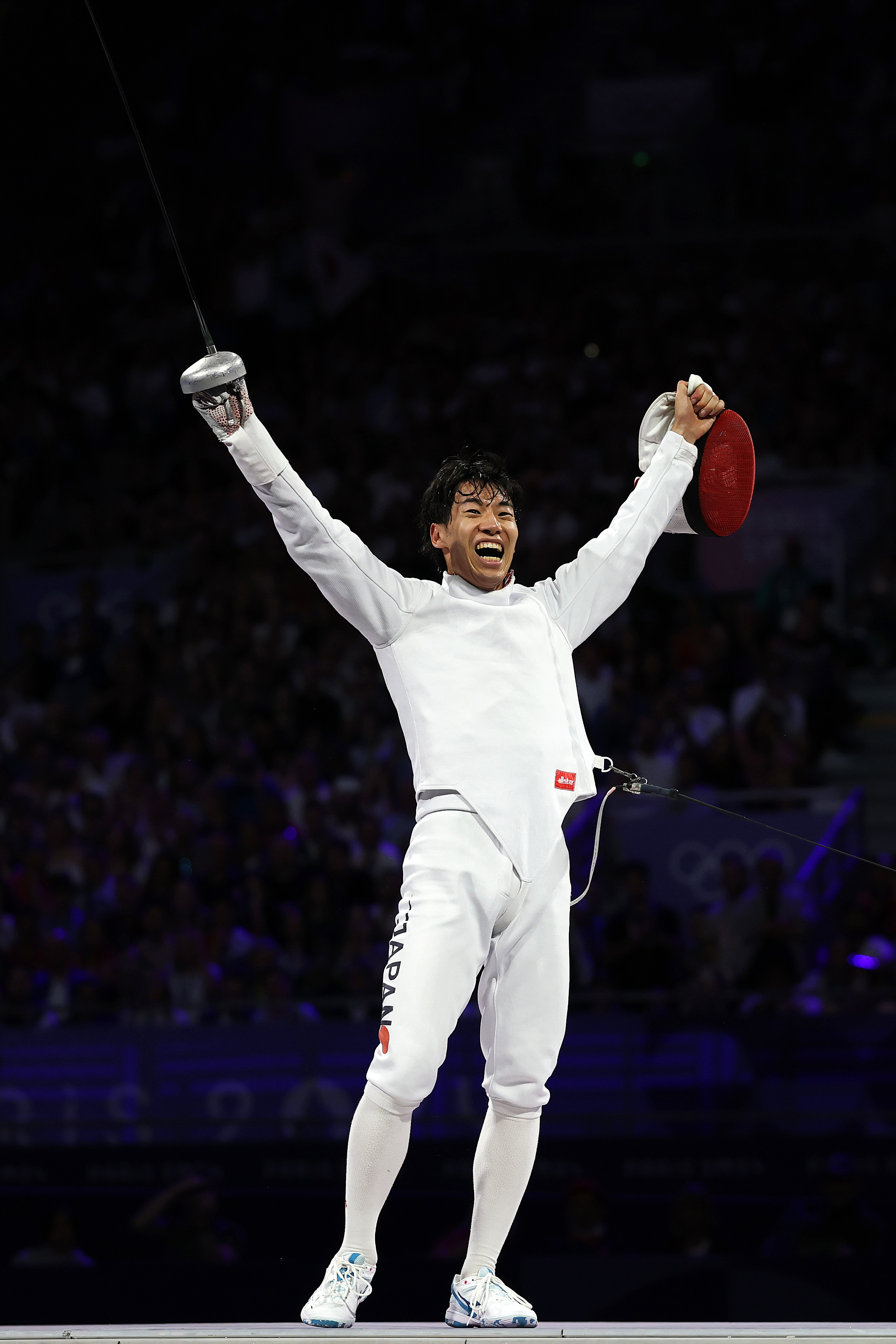A fencer in full gear with the name &quot;Japan&quot; on his uniform lifts his arms in victory, holding his foil and mask, celebrating his win