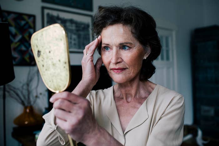 An elderly woman with short, wavy hair examines her reflection in a handheld mirror, touching her forehead with a thoughtful expression