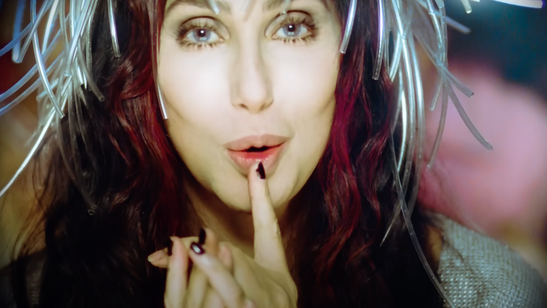 Cher, in a striking headdress of silver strands, holds a finger to her lips in a shushing gesture, her nails painted black