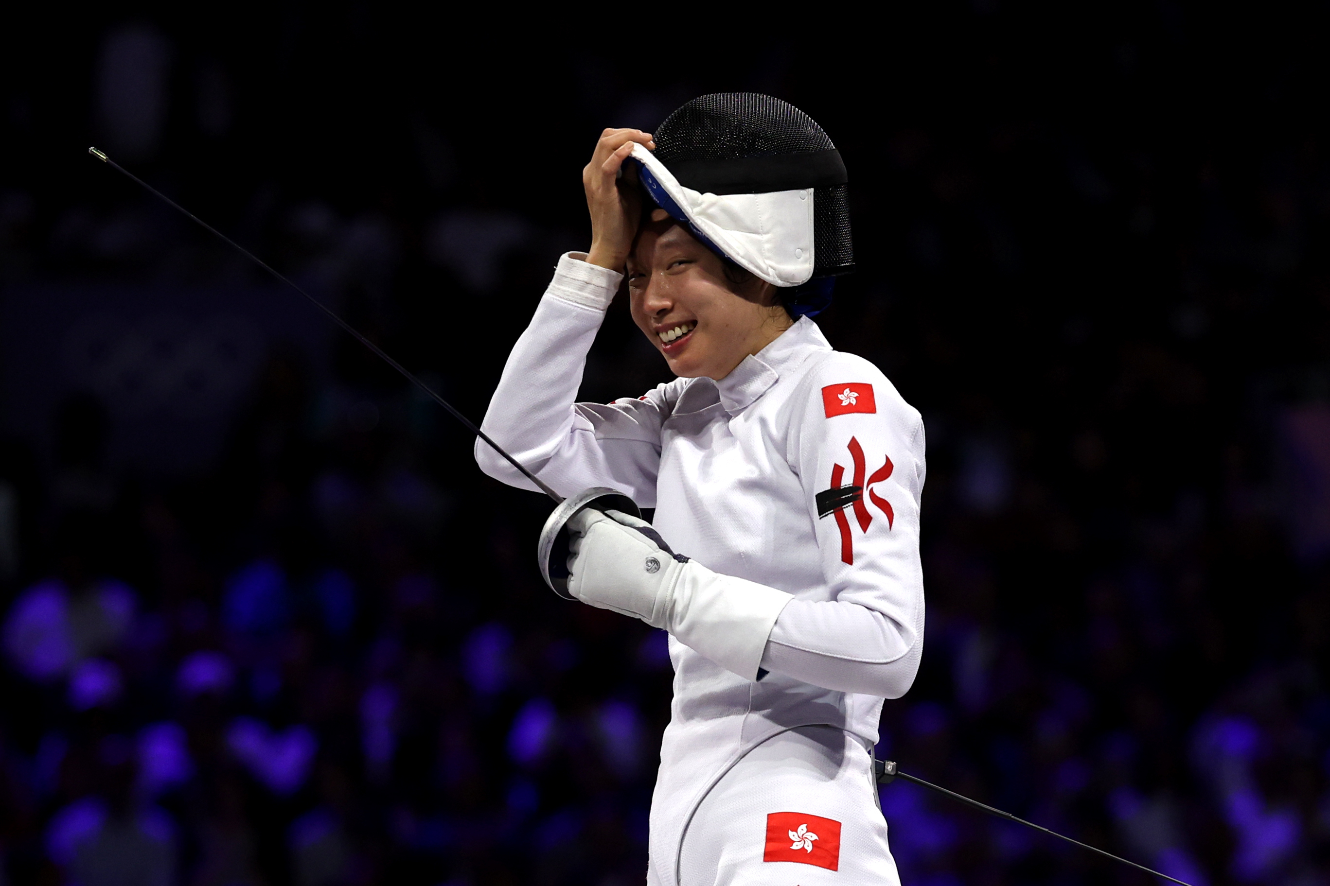 Fencer Vivian Kong removes his mask and smiles after a match. He is wearing a full white fencing uniform adorned with Hong Kong flags