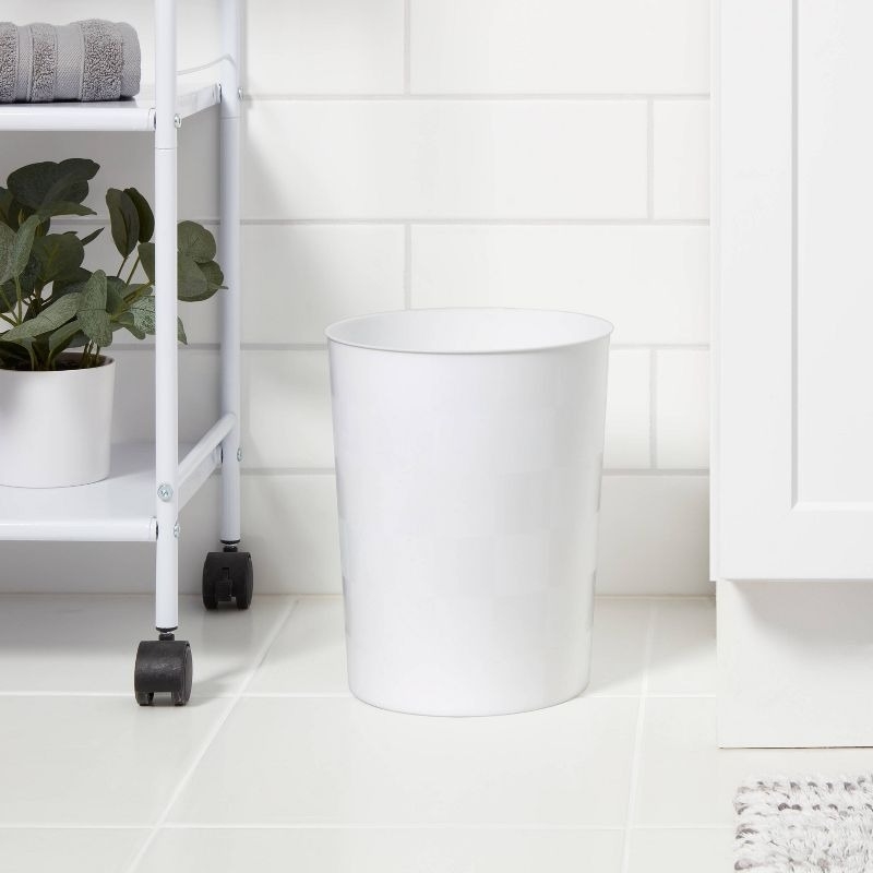 A white trash can is placed in the corner of a bathroom with white tiled walls and floor. A small shelf with a potted plant and towels is beside it